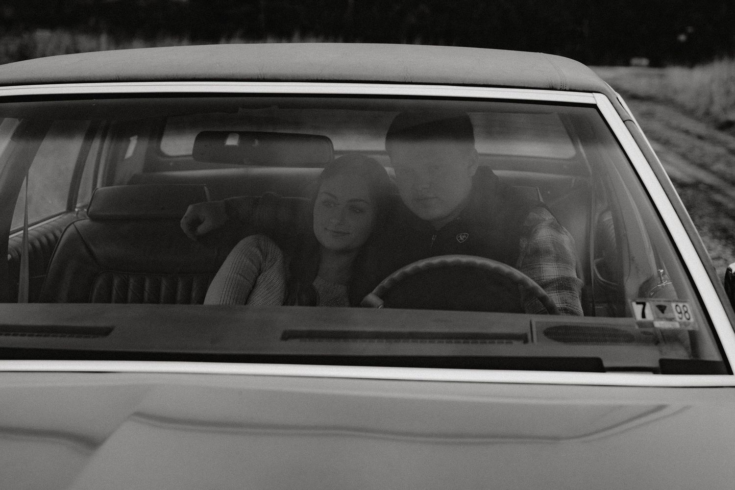 A black and white view through a classic car windshield showing people in the front seat.