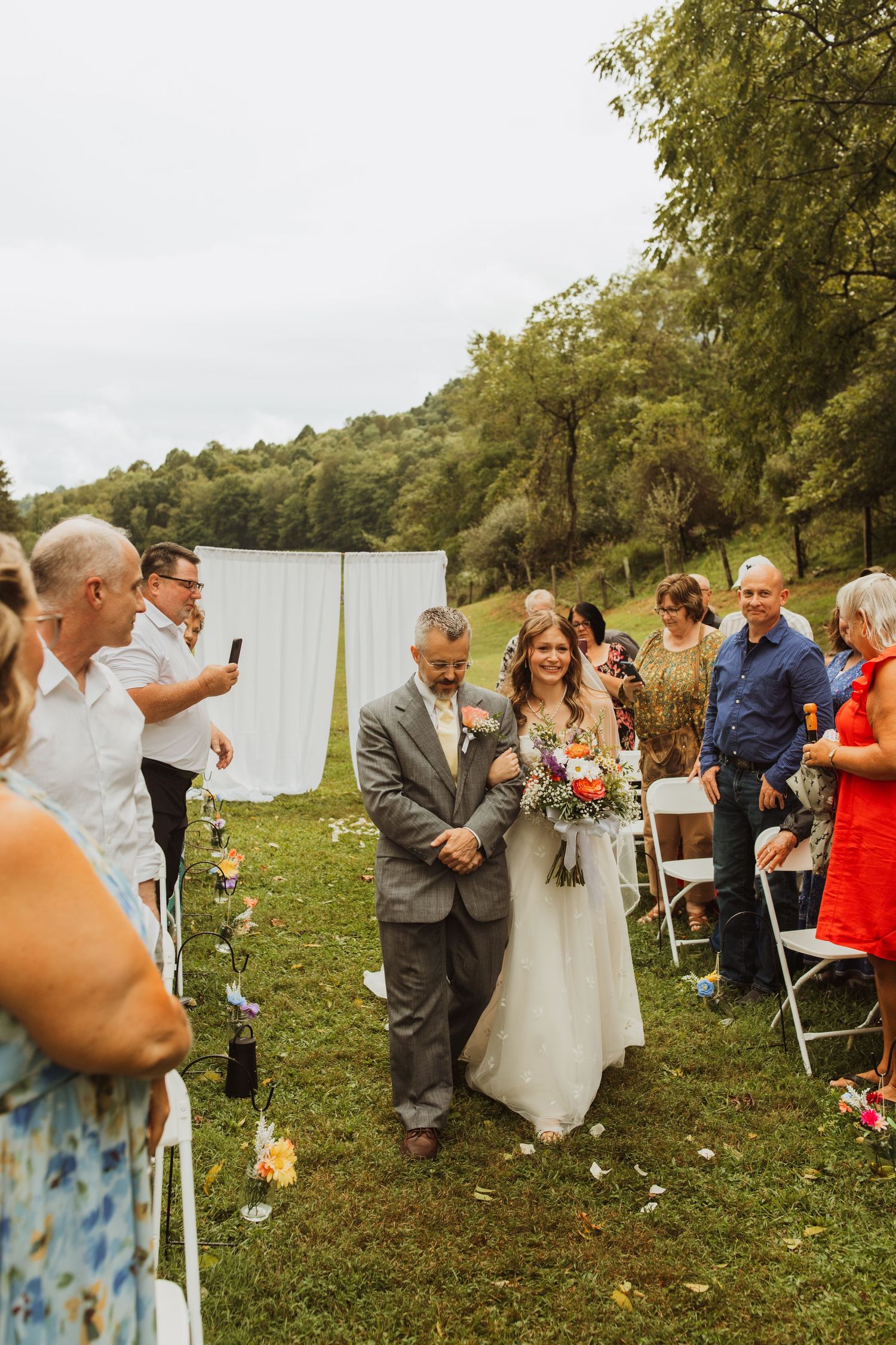 Wedding ceremony processional on a green lawn with white backdrop and folding chairs.