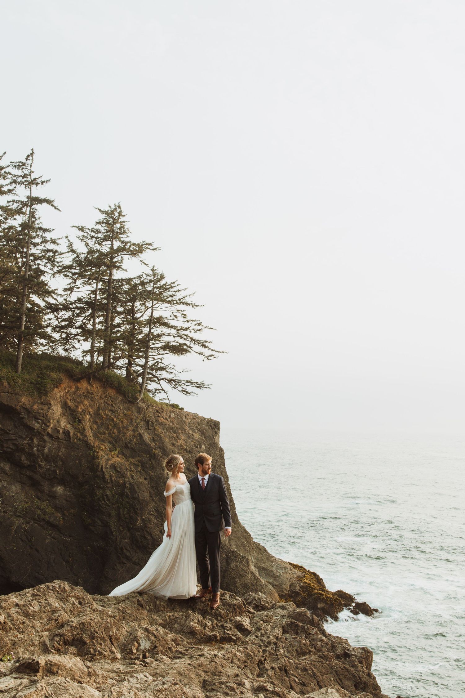 A couple in wedding attire stands on rugged ocean cliffs with evergreen trees and misty waters in background.