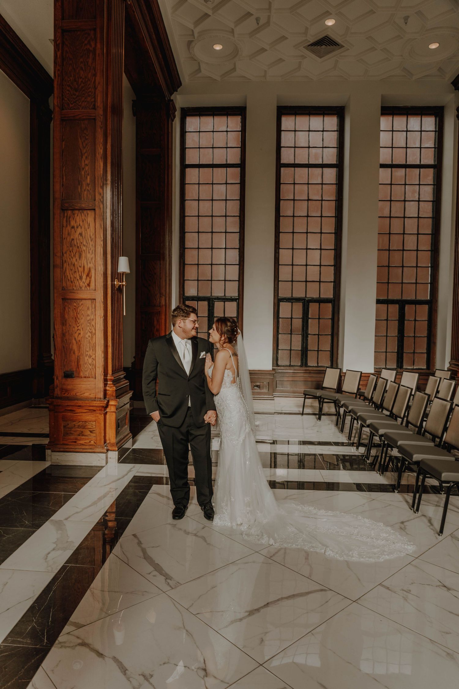 A wedding couple stands in an elegant venue with tall windows and marble floors.