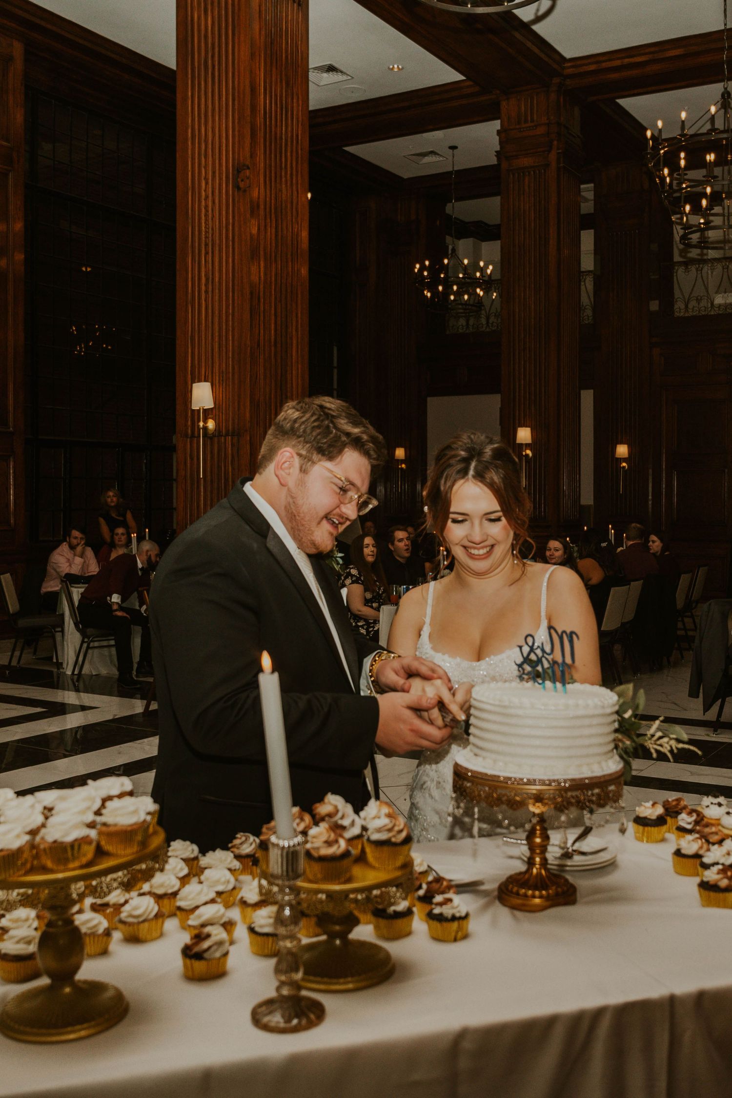 A couple cutting their white wedding cake surrounded by cupcakes at an elegant reception.