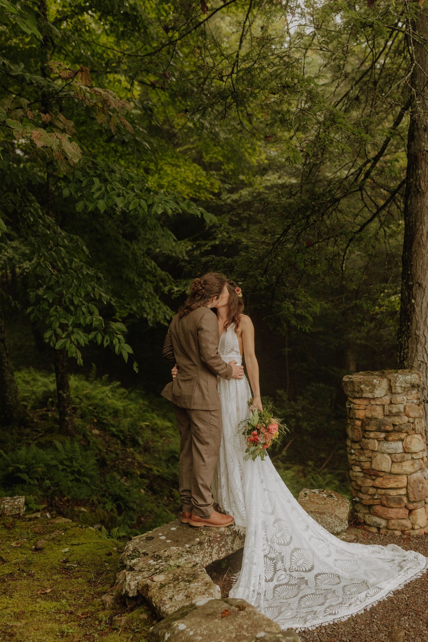 A tender moment shared between newlyweds near a stone pillar in a lush forest setting.