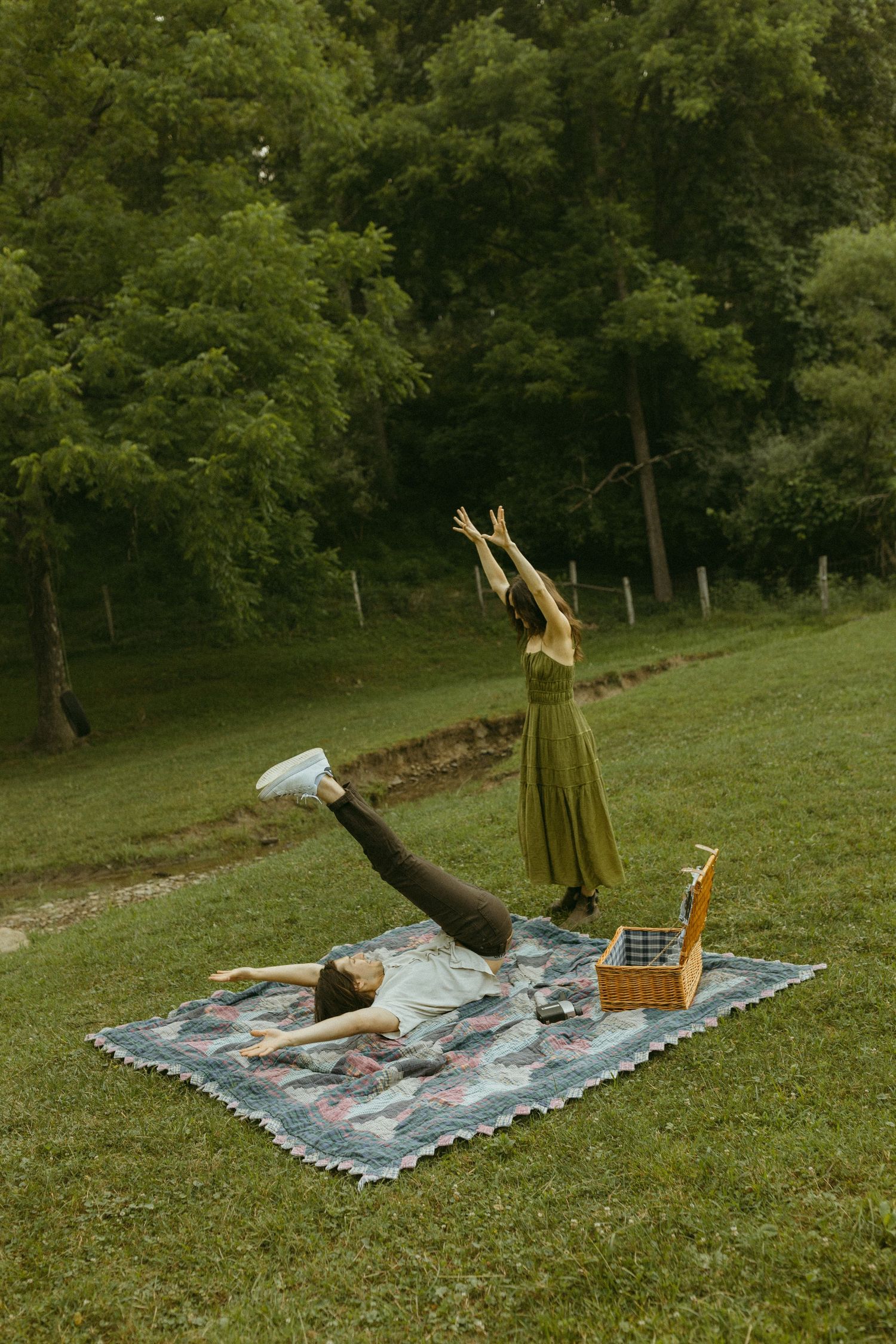 Two people having a playful picnic photoshoot with one striking a dramatic pose on the blanket.