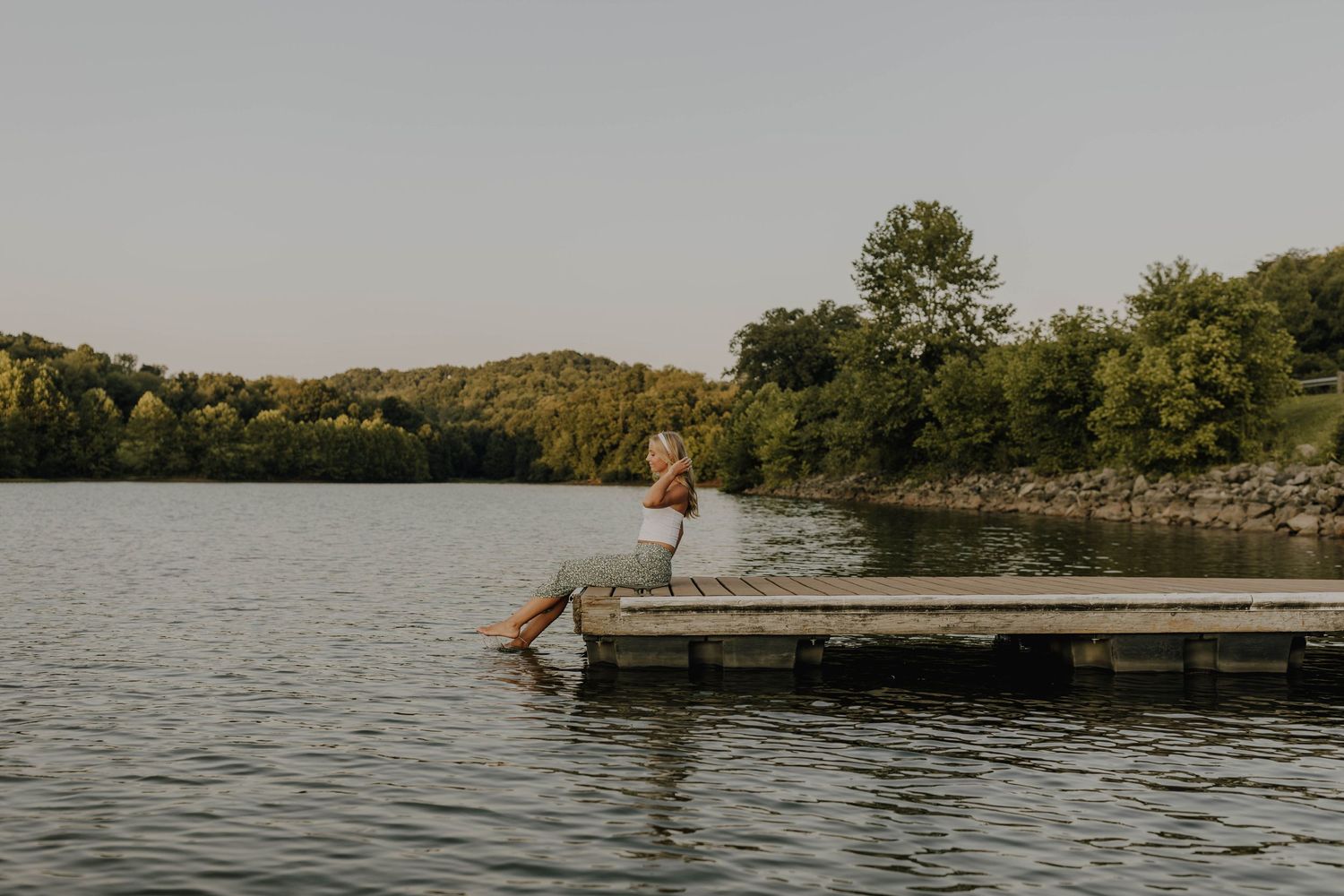 Figure in white dress jumps off wooden dock into serene lake at sunset.