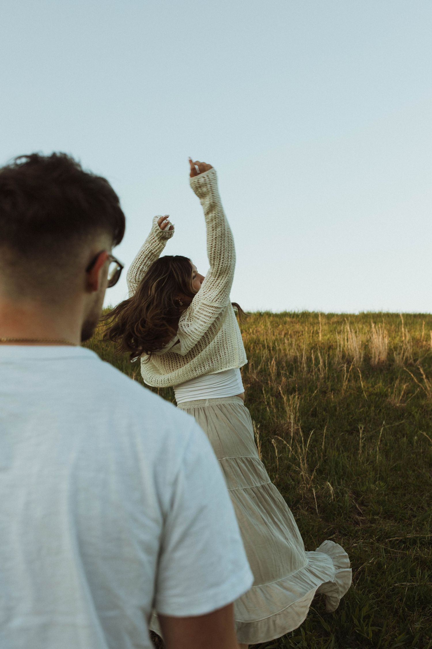 A couple enjoying a playful moment with raised arms in a grassy field at sunset.