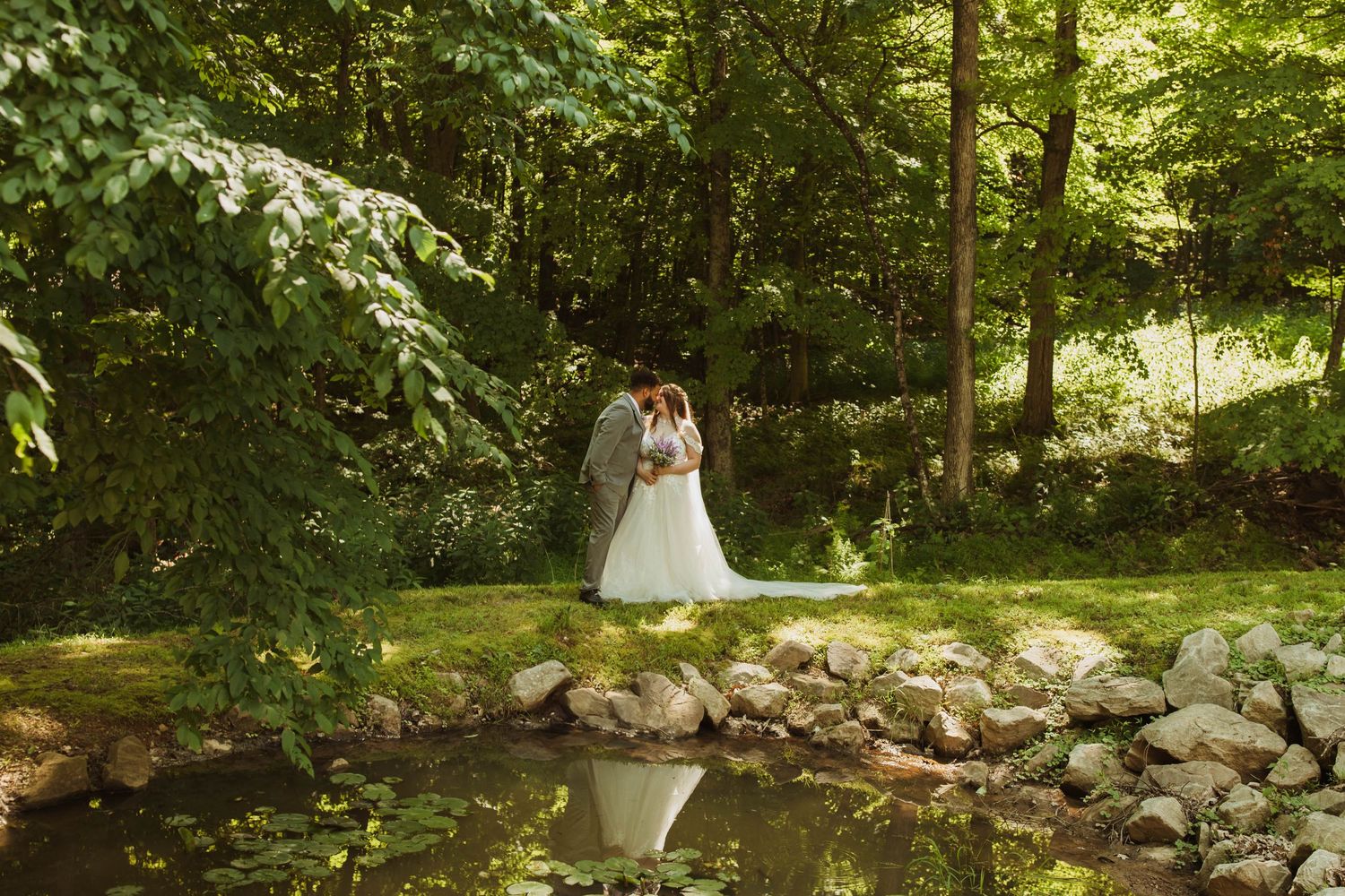 Wedding couple stands together on stone rocks near a reflective pond in a serene woodland setting.