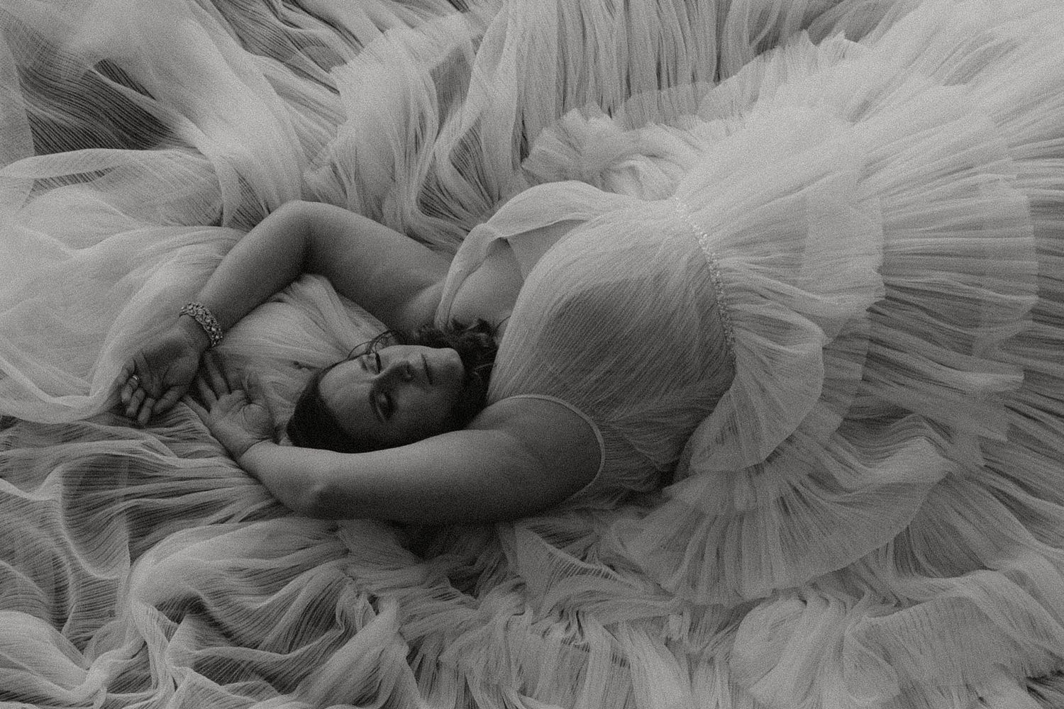 Black and white photo of a person nestled in layers of flowing white tulle fabric.