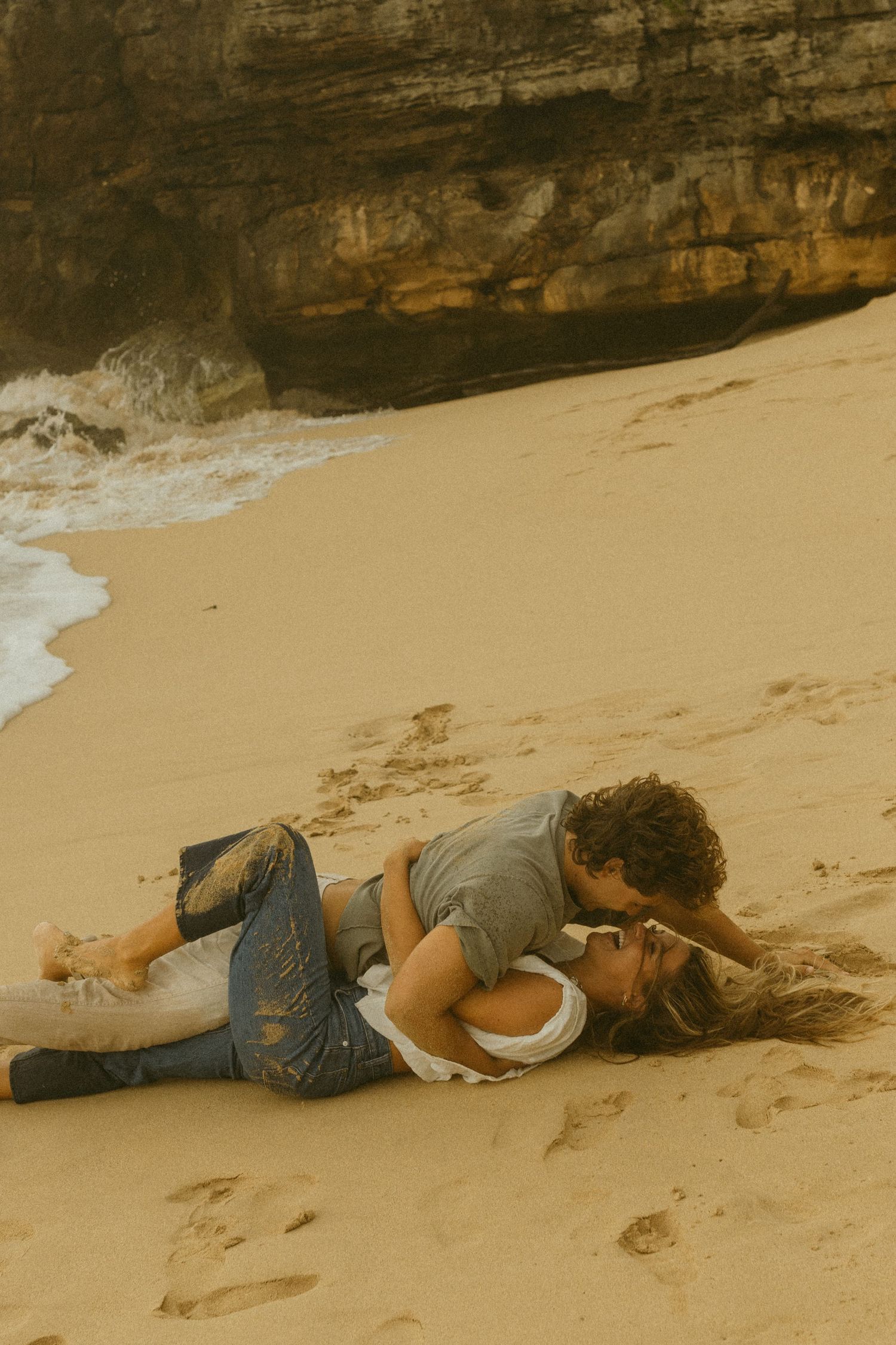 Couple lying close together on sandy beach near rocky cliffs.