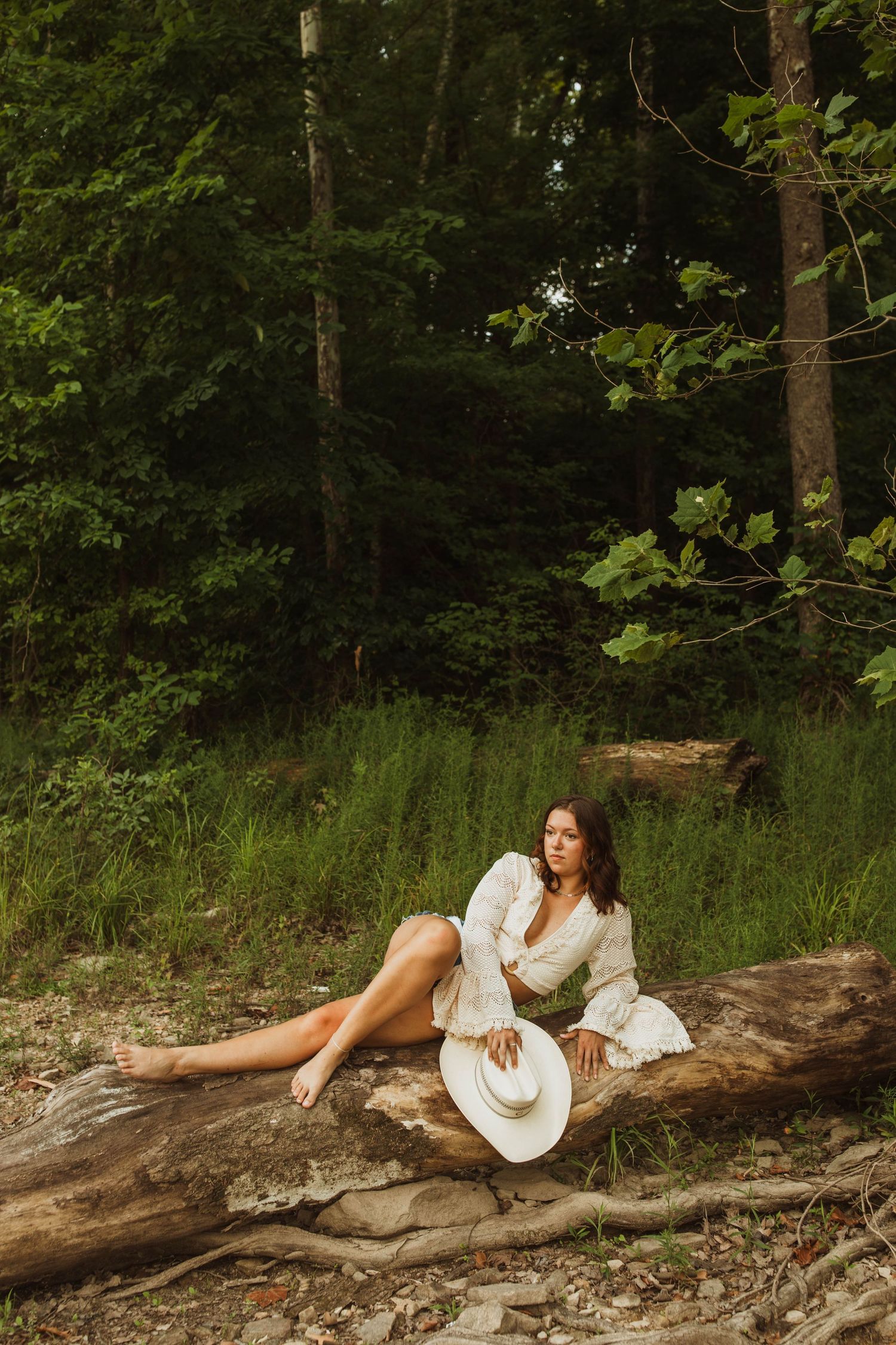 Person in white dress reclines on fallen log in wooded forest setting.