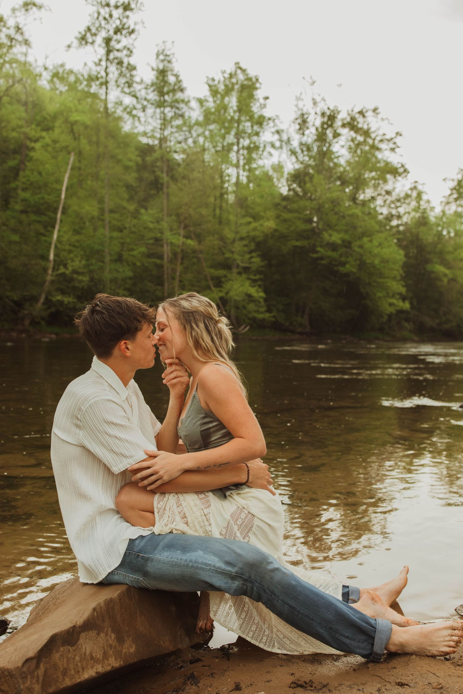 Two people share a tender moment while sitting on rocks beside a flowing river.