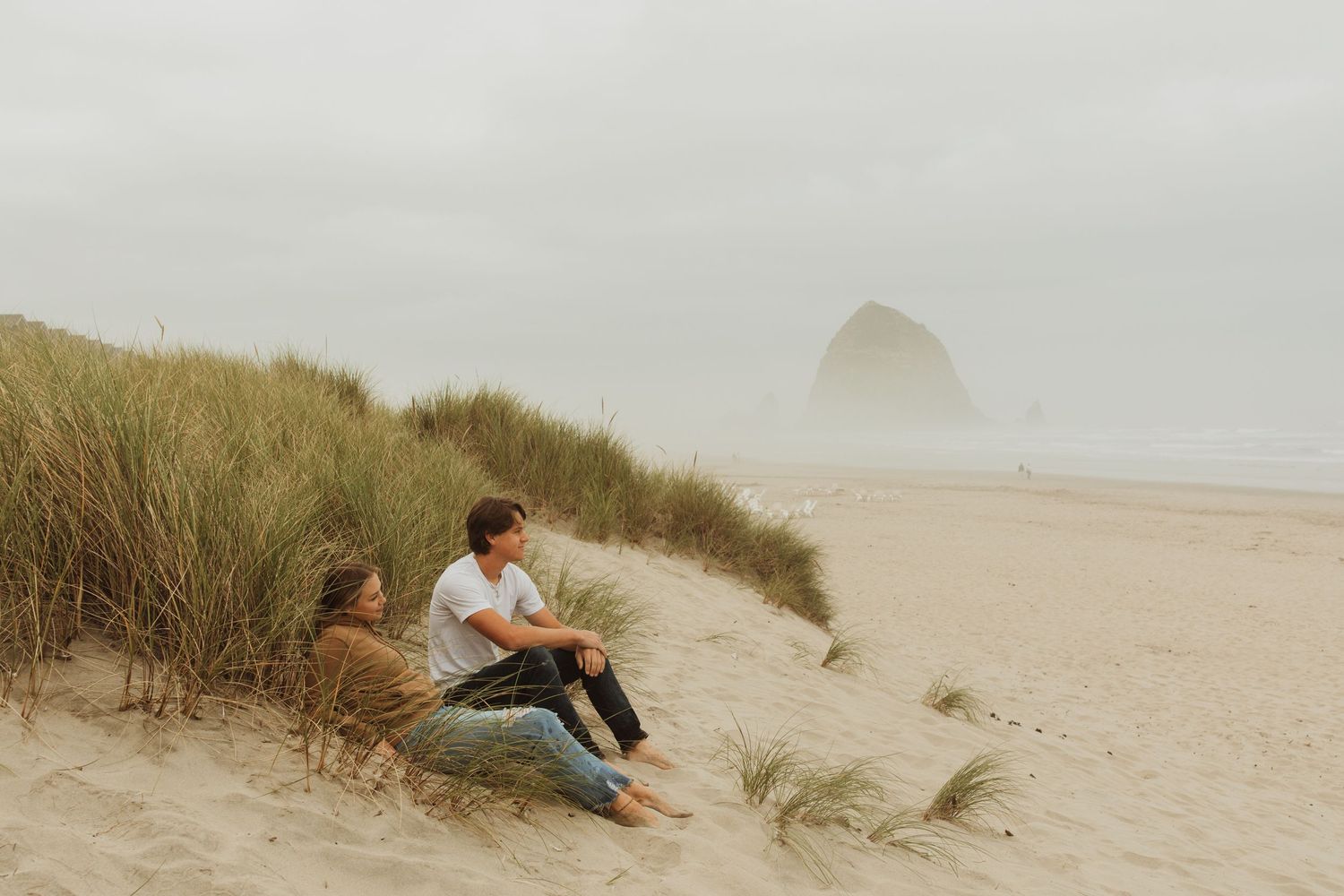 Couple relaxing on sand dunes overlooking a foggy beach with iconic Haystack Rock visible in the background.