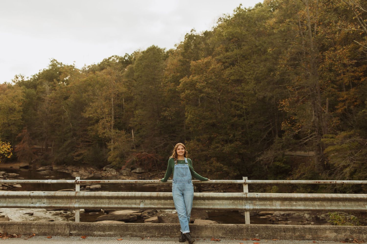 Casual pose on bridge railing with overalls against backdrop of autumn lake and forest.