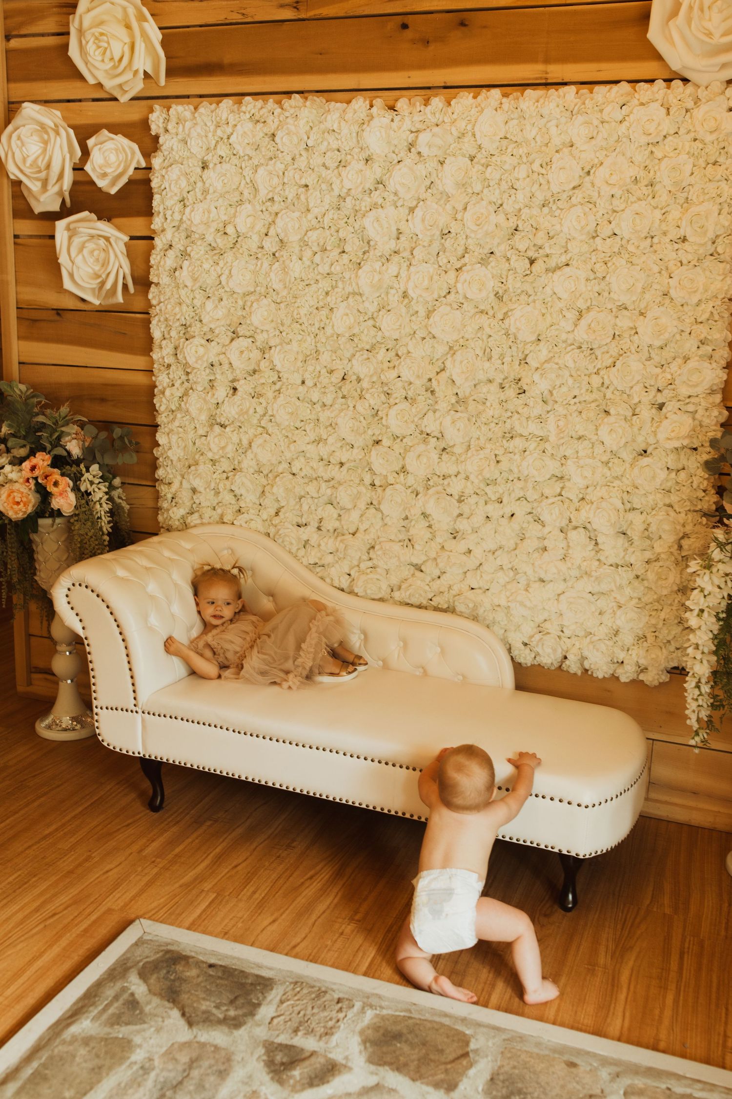 White floral backdrop with cream chaise lounge and baby decor in rustic wooden room.