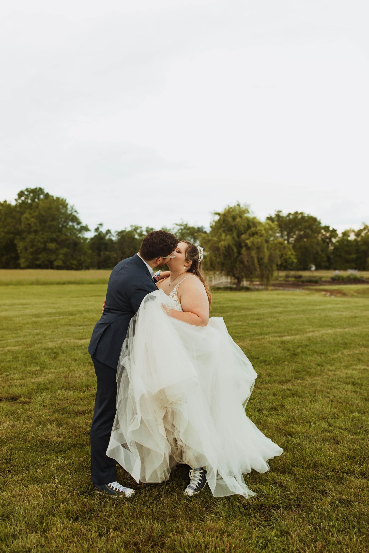 A bride and groom embrace and kiss while standing in a grassy field.
