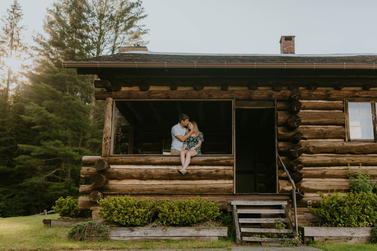 A rustic log cabin with a front porch nestled among evergreen trees at sunset.