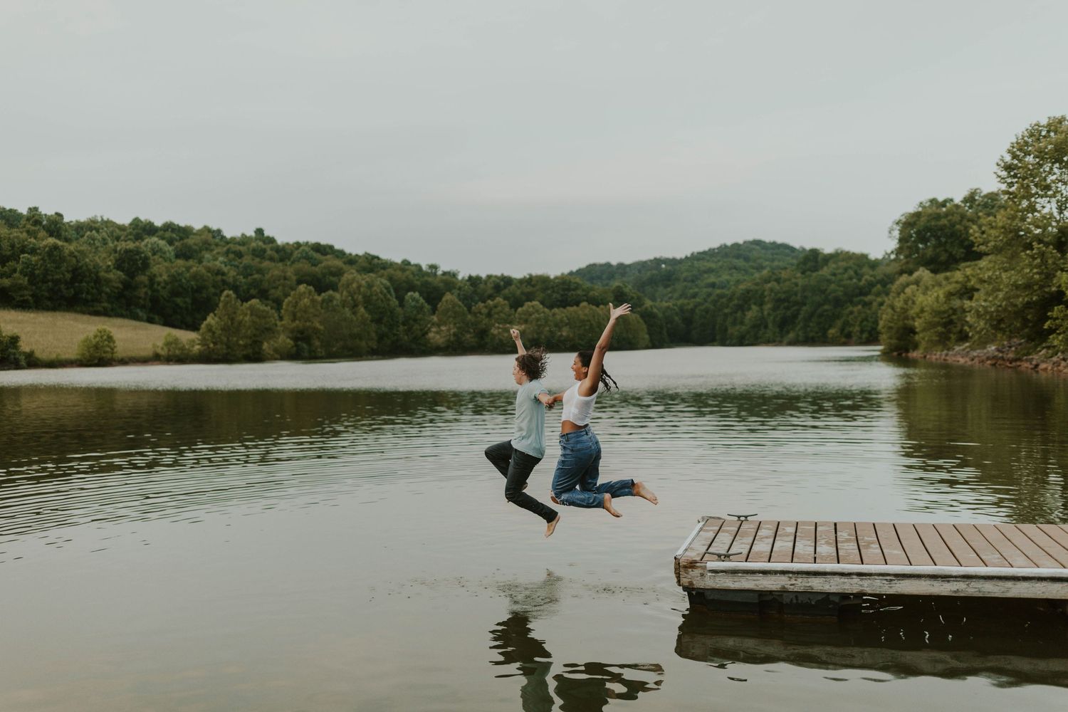 Two people jumping in unison off a wooden dock into a tranquil lake during sunset.