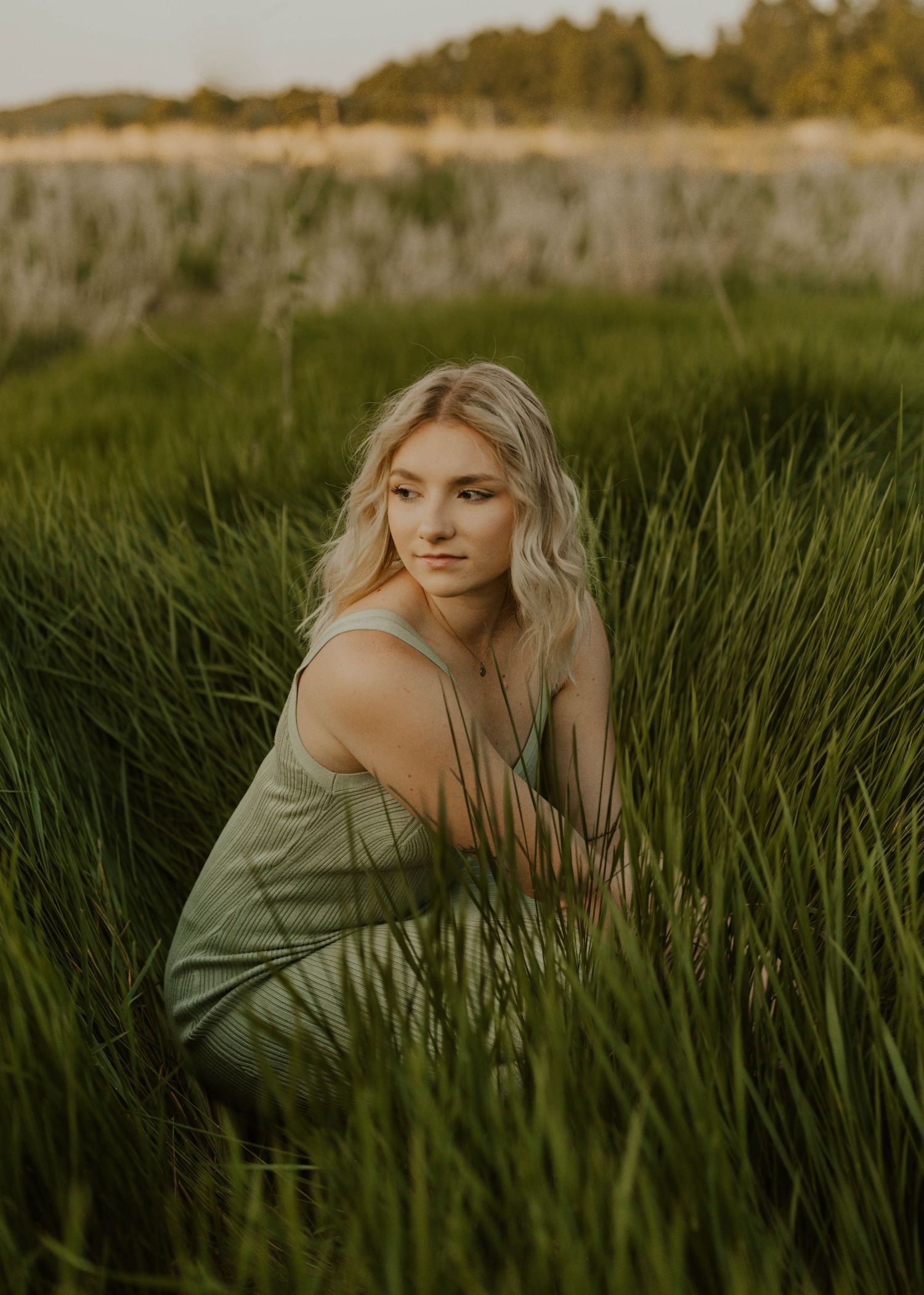 Portrait of person in olive dress sitting among tall grass at sunset.