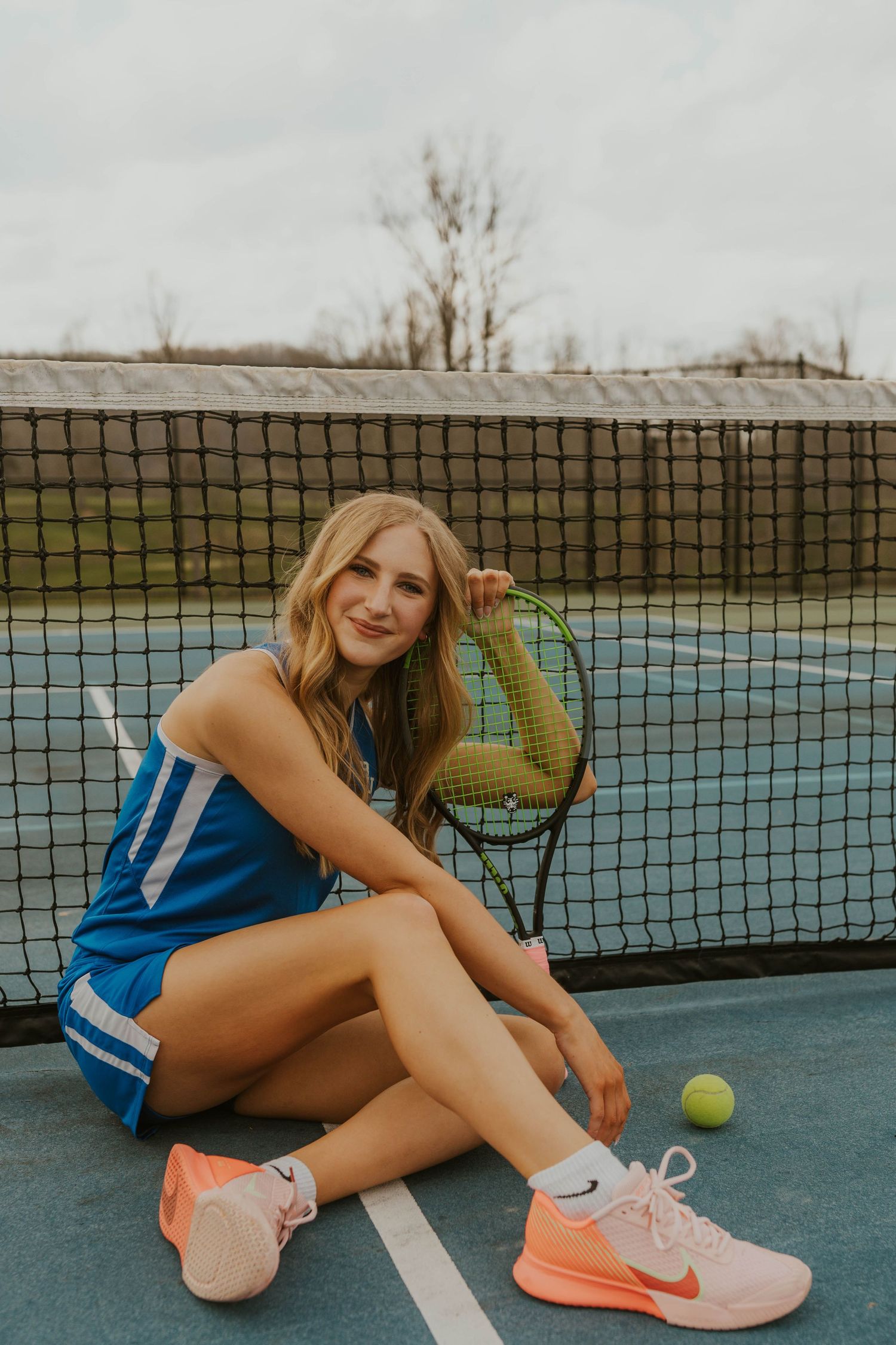 A tennis player sits on a court with a racket and ball wearing blue athletic gear.