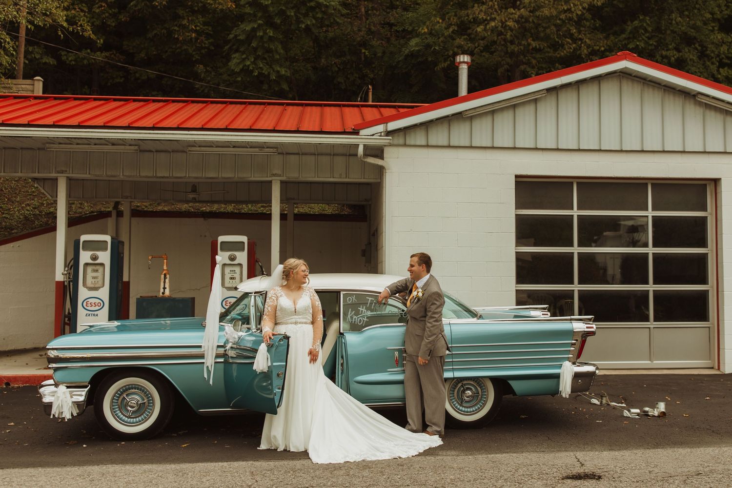 A wedding couple poses with a retro turquoise car at a vintage gas station.