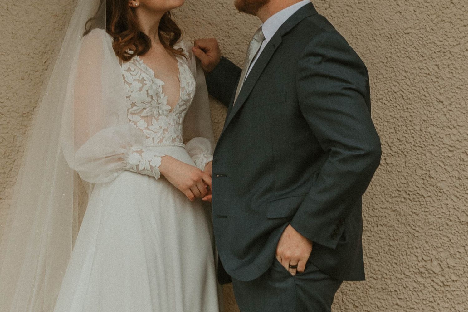 Intimate wedding portrait showing details of bride's lace dress and groom's dark suit against neutral wall.