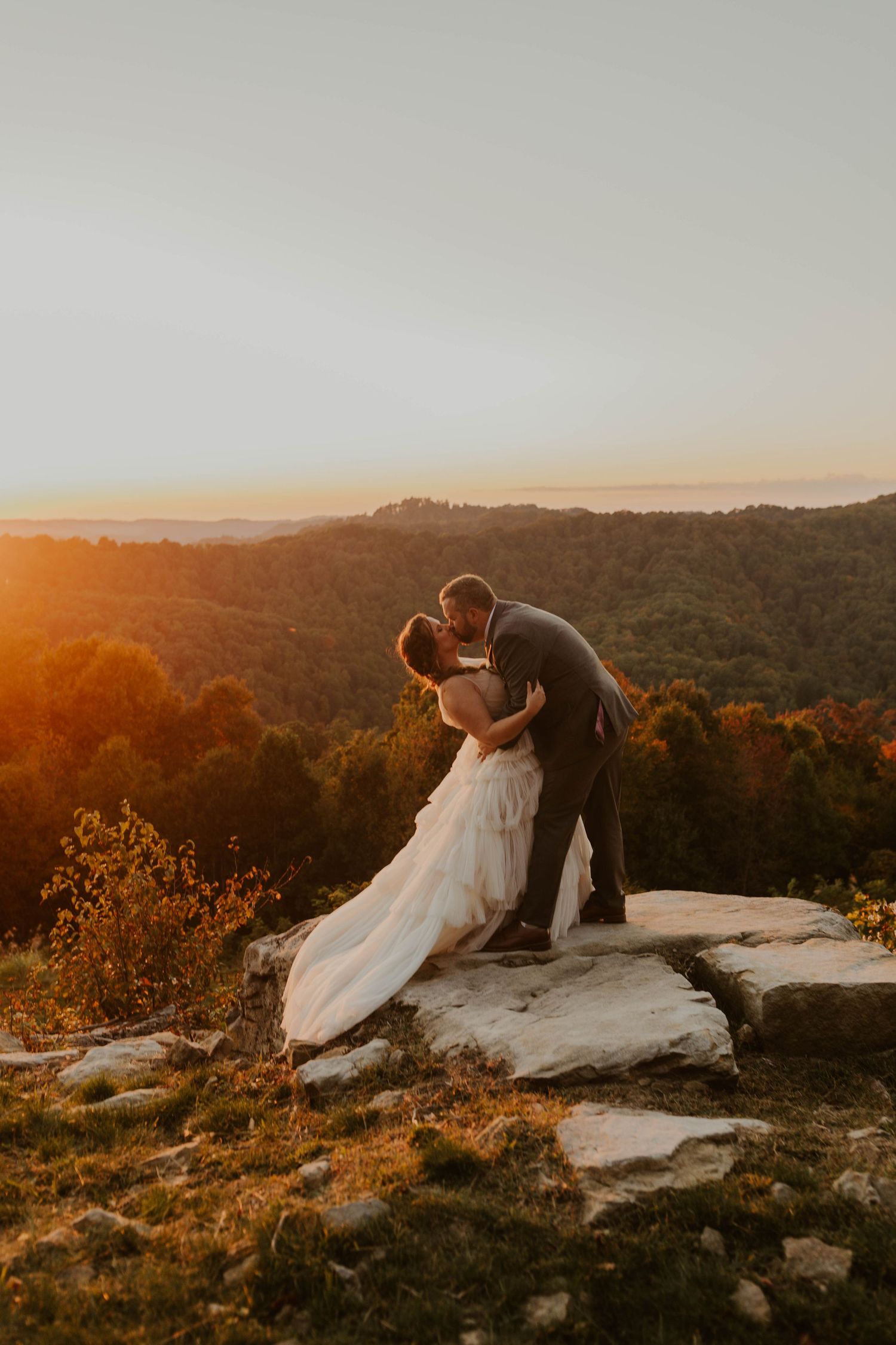 Wedding couple shares intimate moment on cliff edge during golden hour sunset.