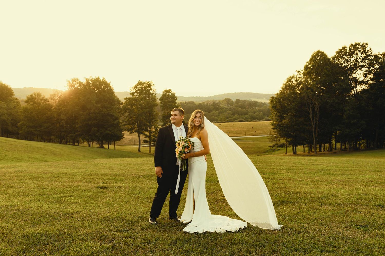 Sunset wedding portrait in a golden field with flowing veil and rolling hills in background.