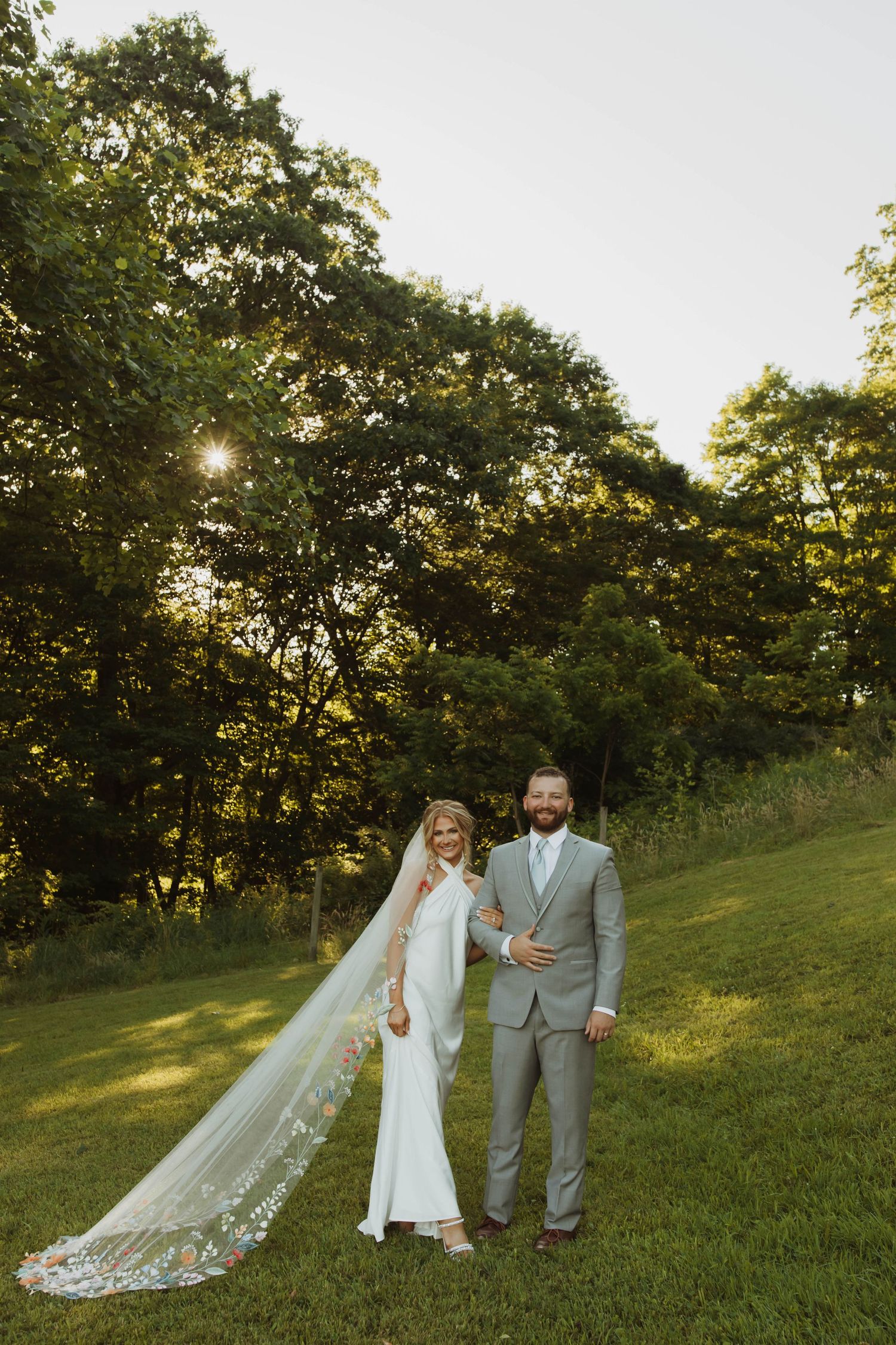 A sunset wedding portrait with a long flowing veil against a tree-lined hillside.