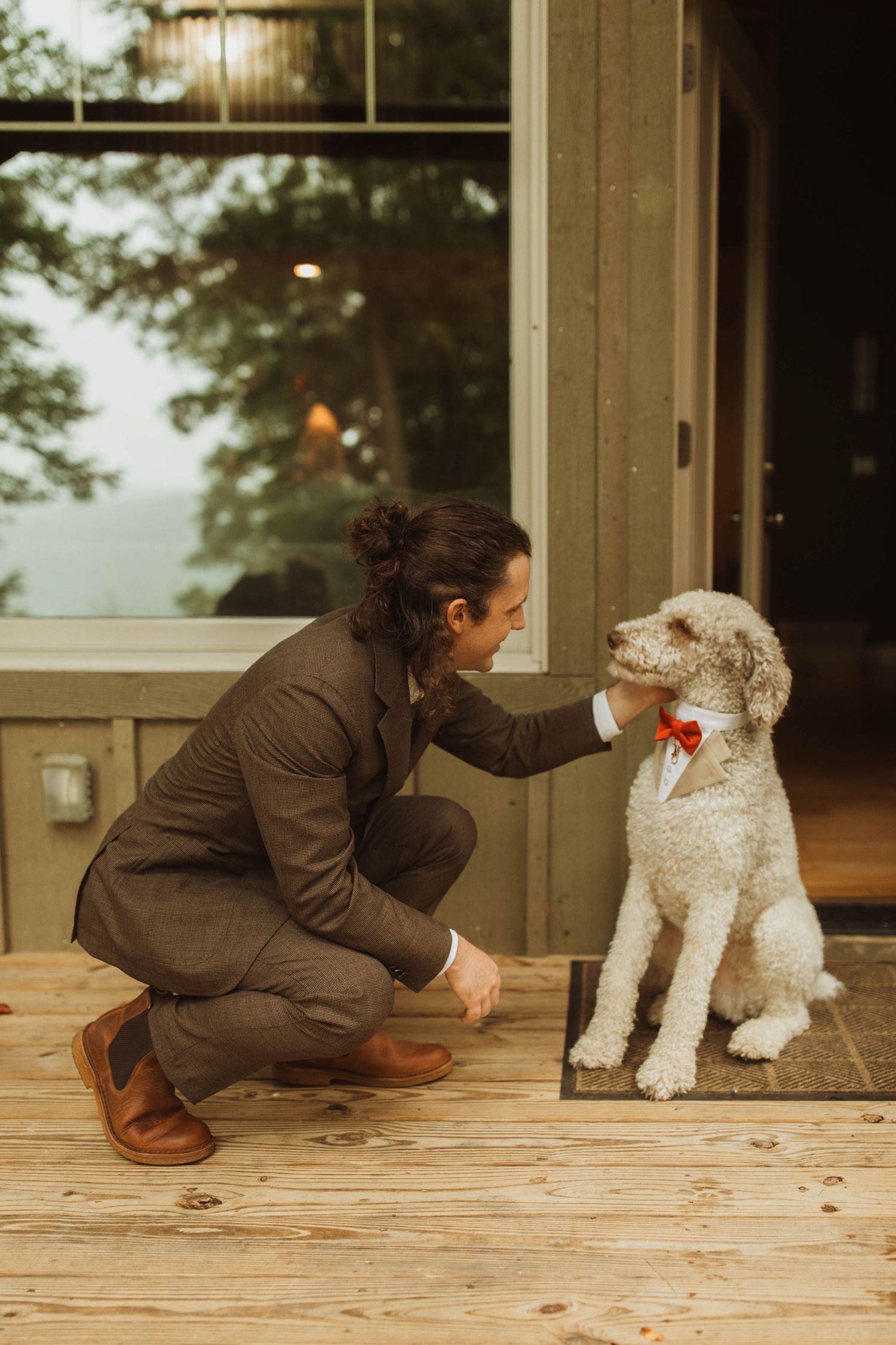 A person in brown attire crouches to pet a fluffy white doodle dog wearing a bow tie.