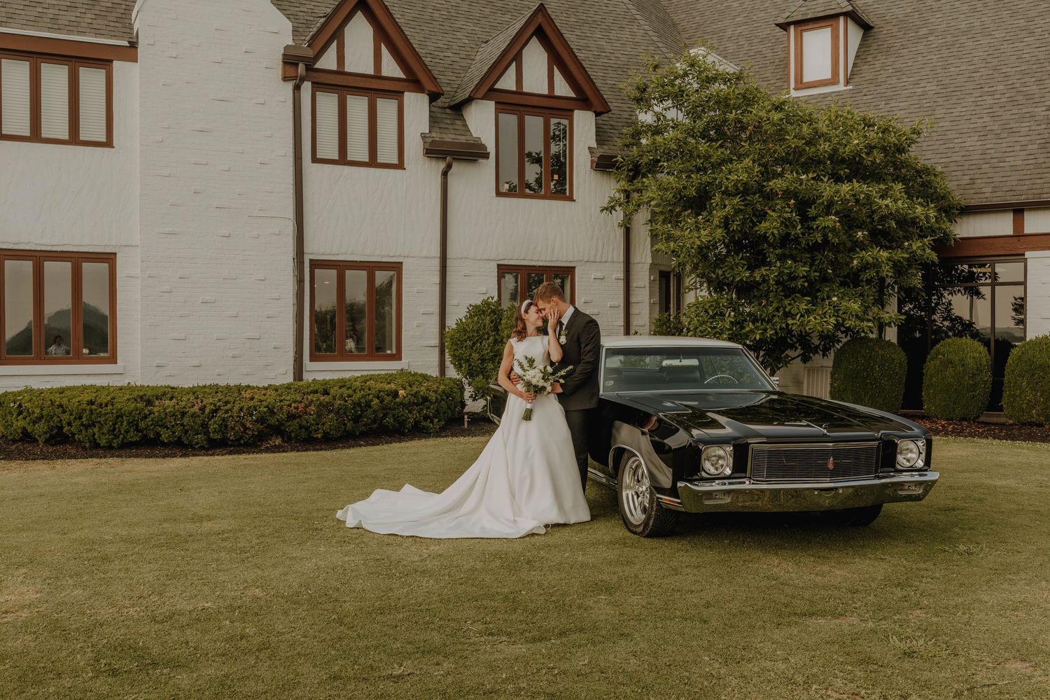 Classic wedding portrait with vintage car and Tudor-style house in background.