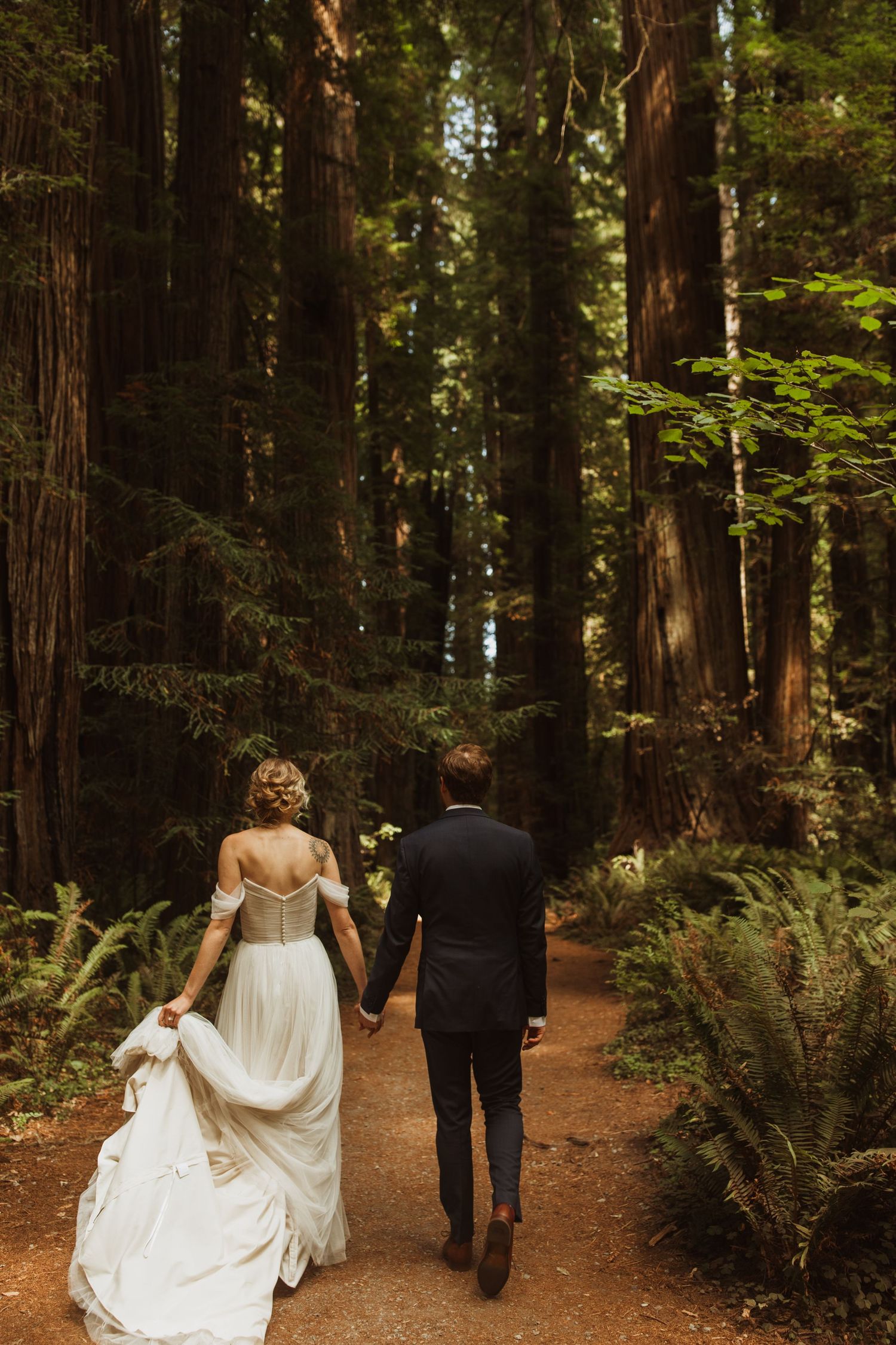 A couple walks hand in hand down a forest path surrounded by towering redwood trees.