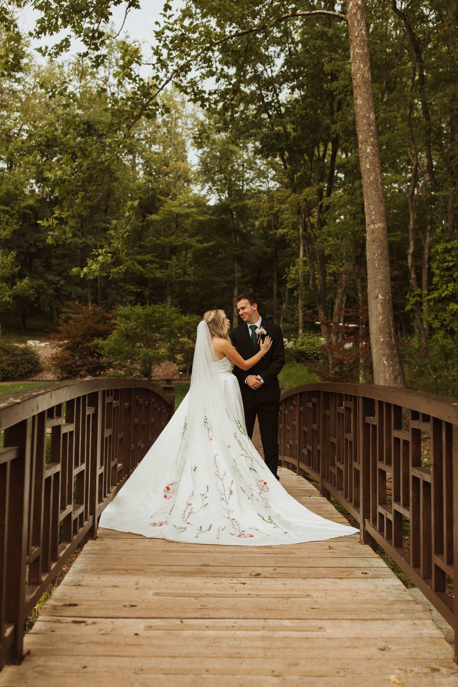 Couple in wedding attire posing on wooden bridge in forest setting.