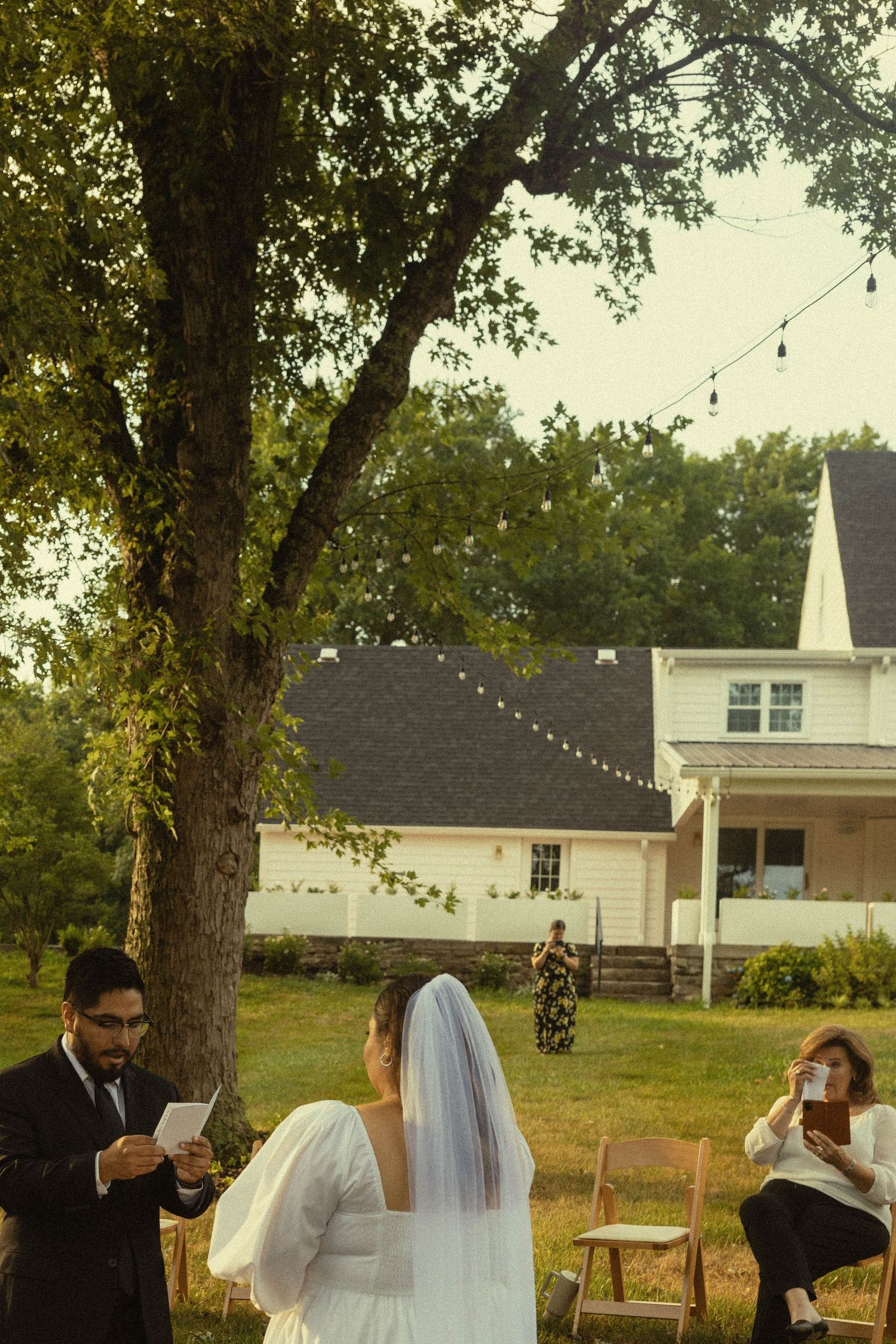 An outdoor wedding ceremony takes place in front of a white country house with string lights.