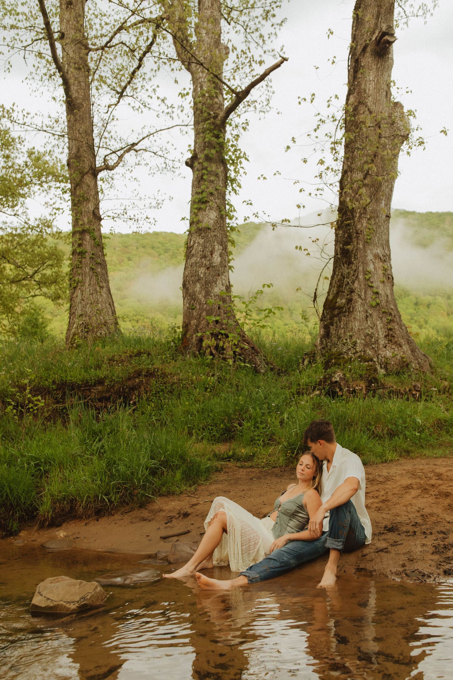 Two people share a peaceful moment sitting by the water's edge near three large trees.
