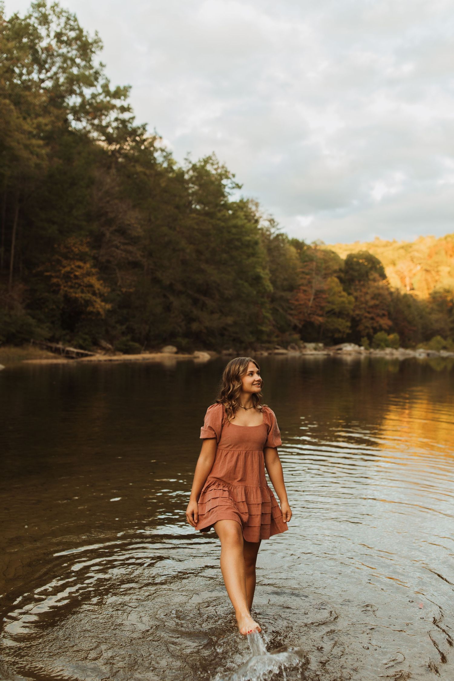 Person in dusty rose dress wading in lake water with golden sunlit forest backdrop.