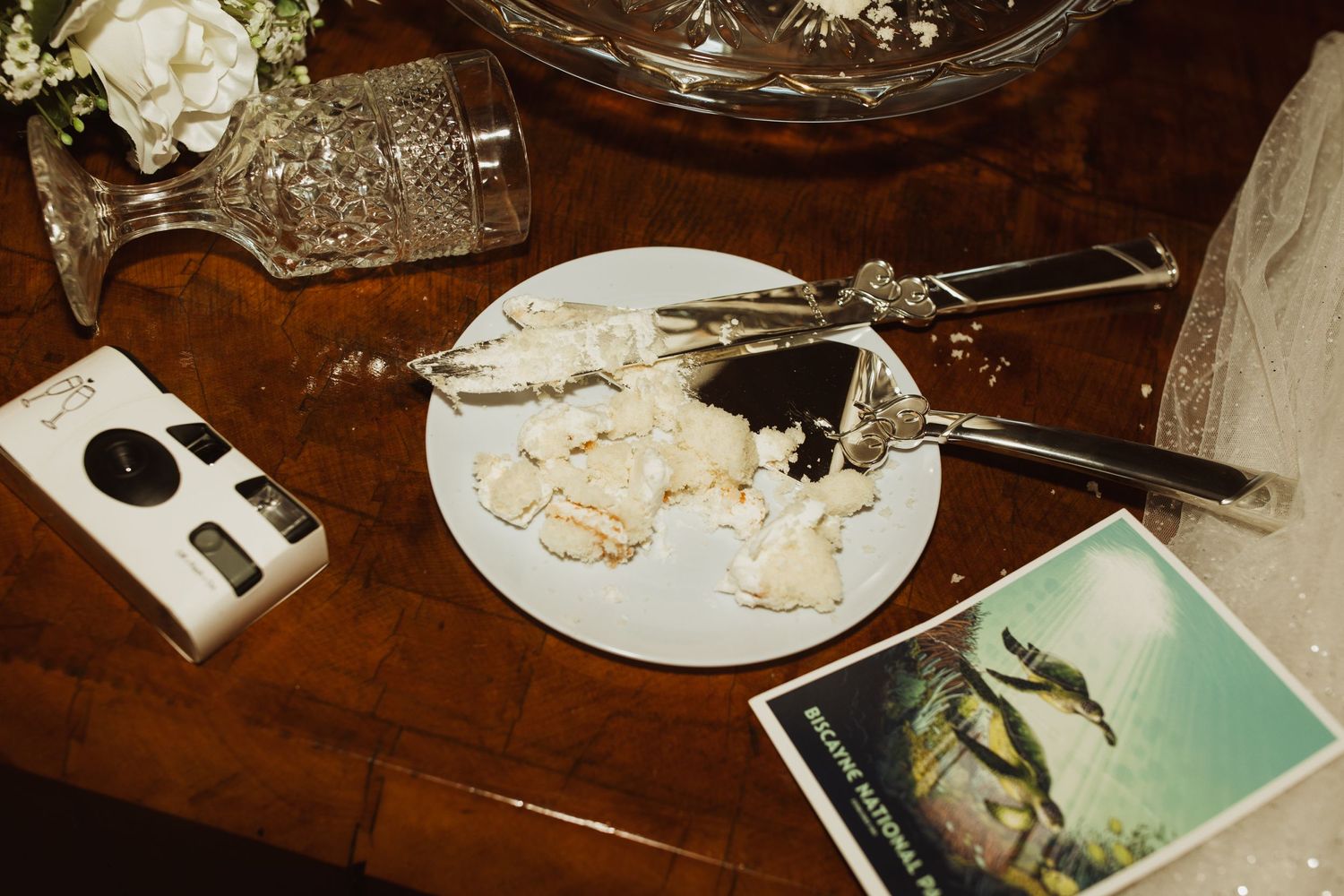 Wedding reception table detail with partially eaten cake and vintage camera on wooden surface.