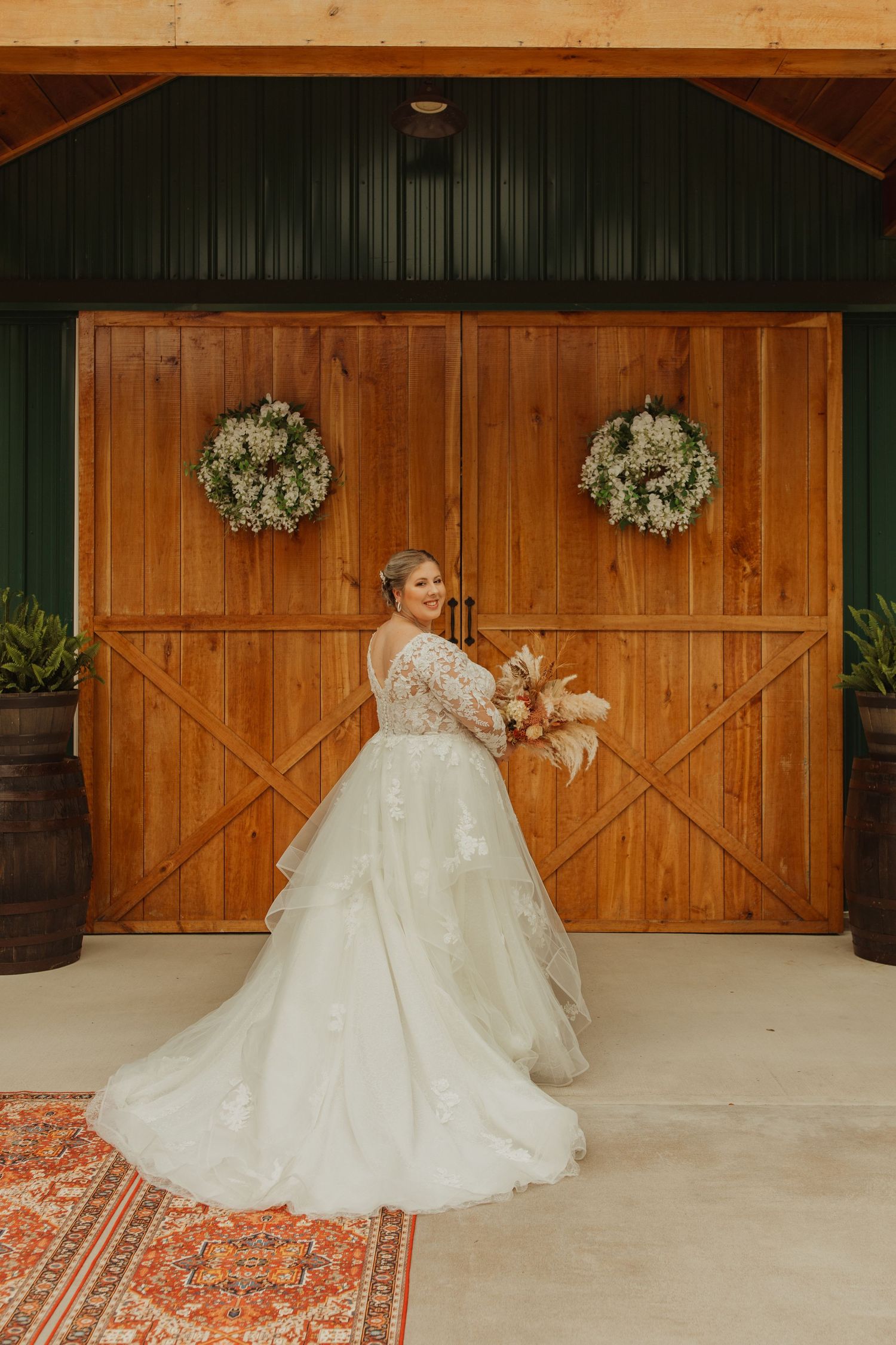 Bride in lace gown poses in front of rustic barn doors decorated with floral wreaths.