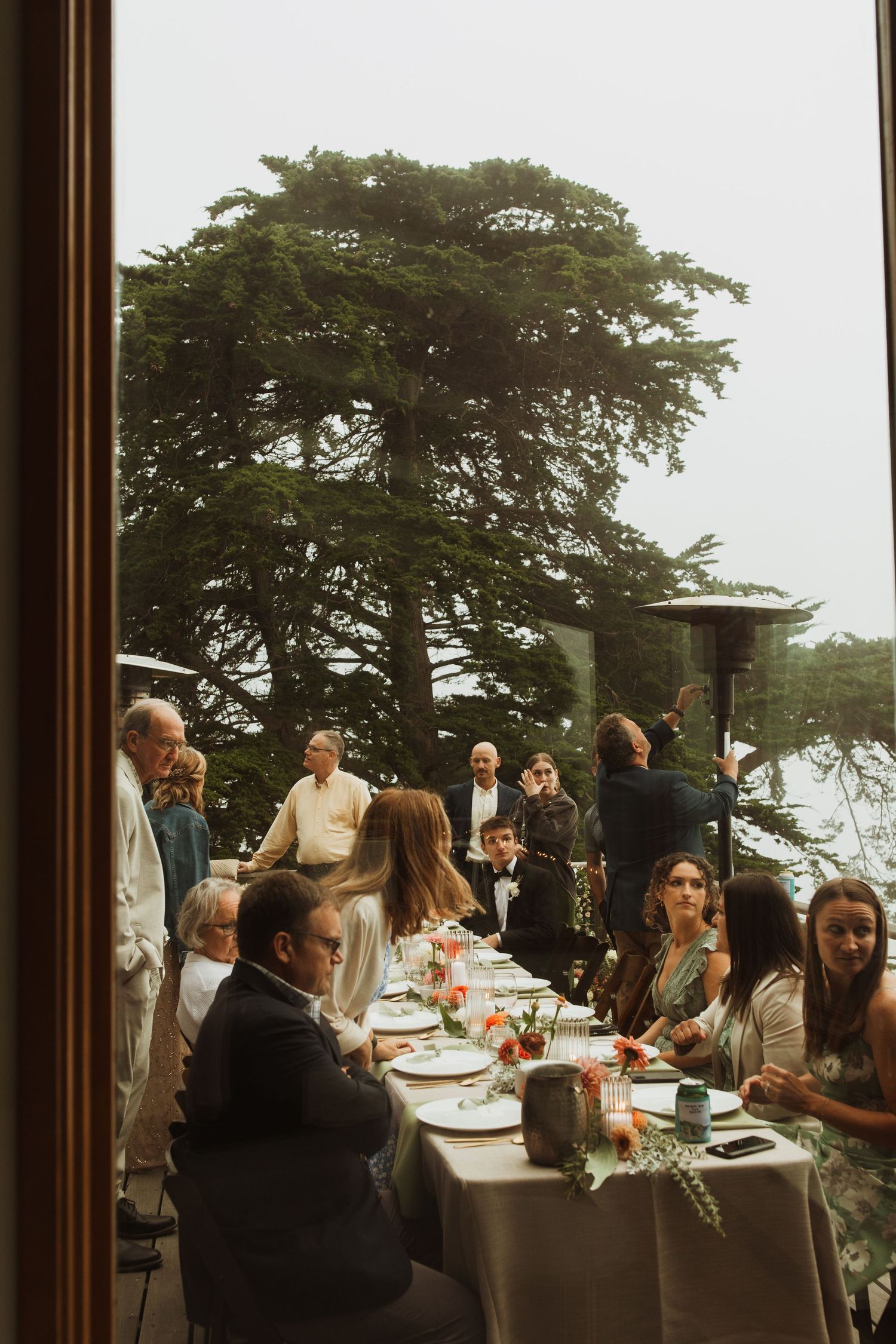 Wedding guests gather around tables on an outdoor deck surrounded by tall pine trees.