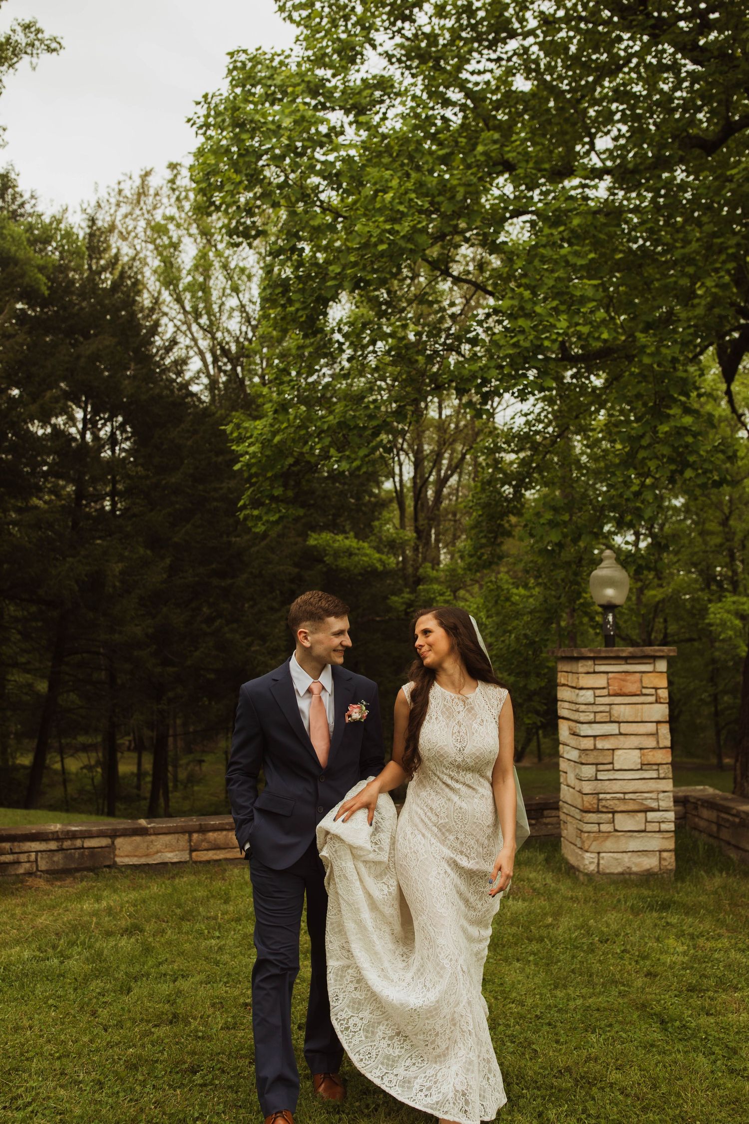 A romantic outdoor wedding scene with a couple walking on grass with a brick pillar and trees in the background.
