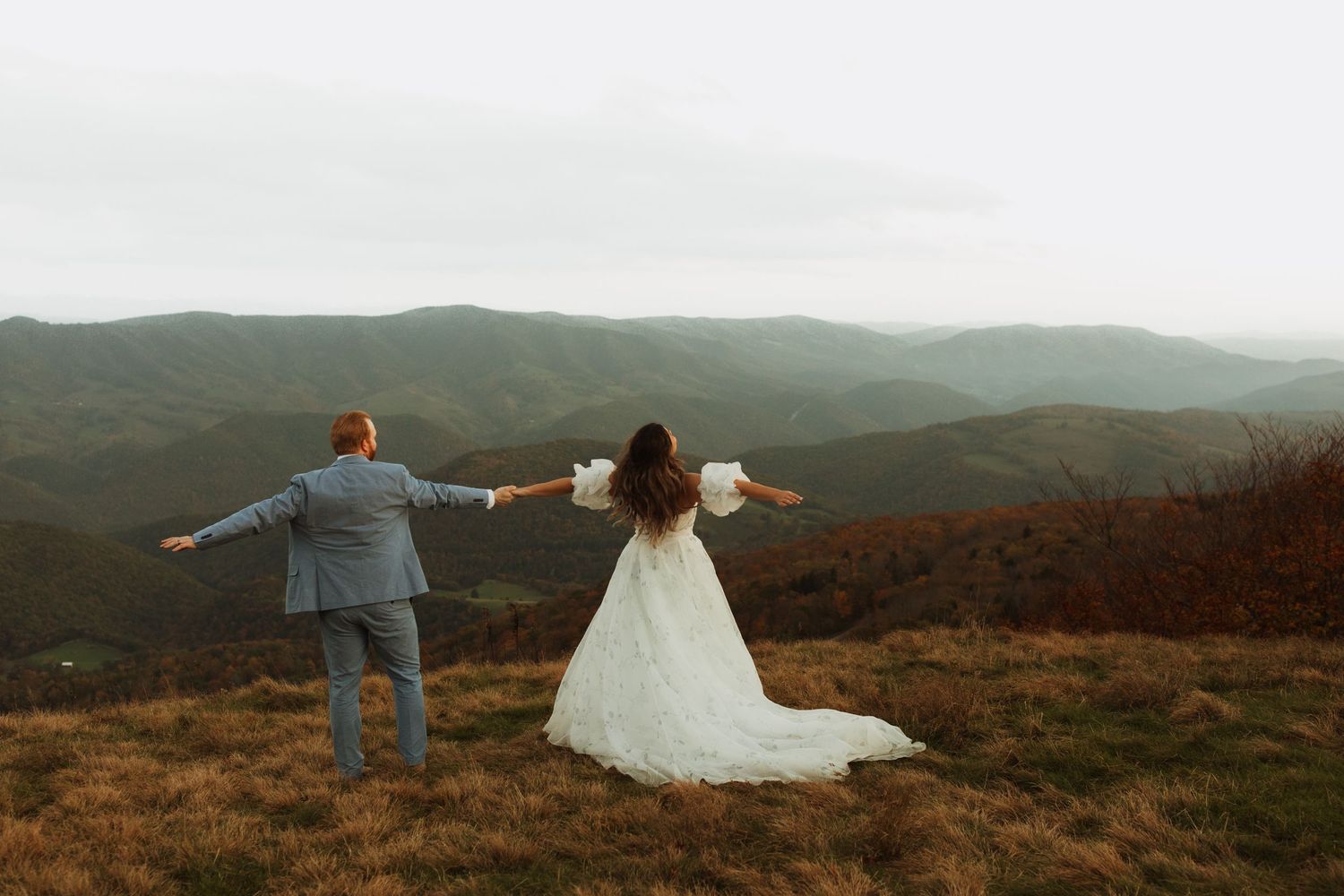 Wedding couple stands with arms outstretched on a mountain ridge overlooking rolling hills.
