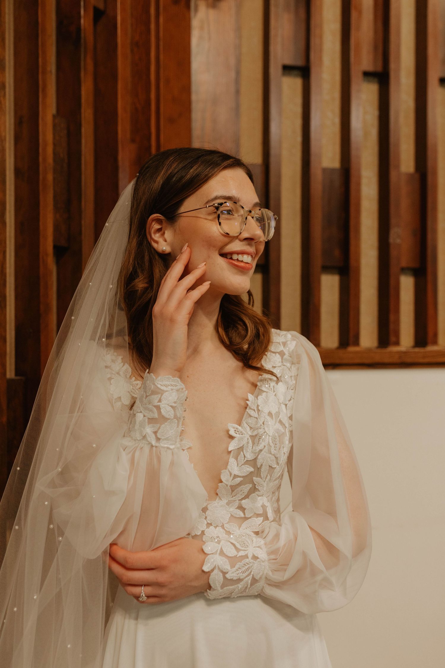 Bride in delicate lace gown and veil smiling while touching her face.