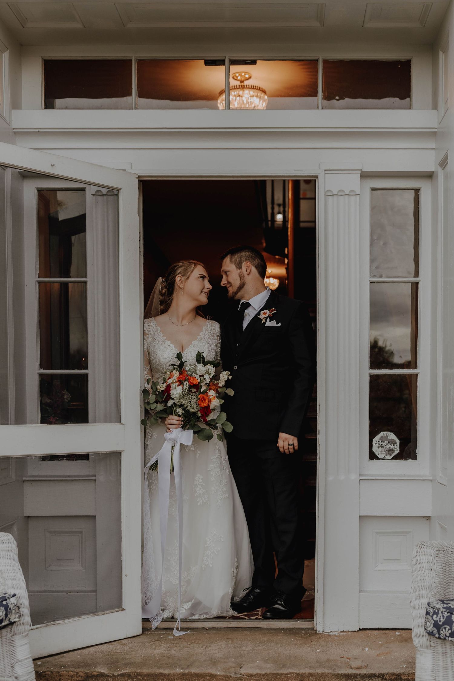 A couple stands in an elegant doorway on their wedding day with bright natural lighting and a romantic bouquet.