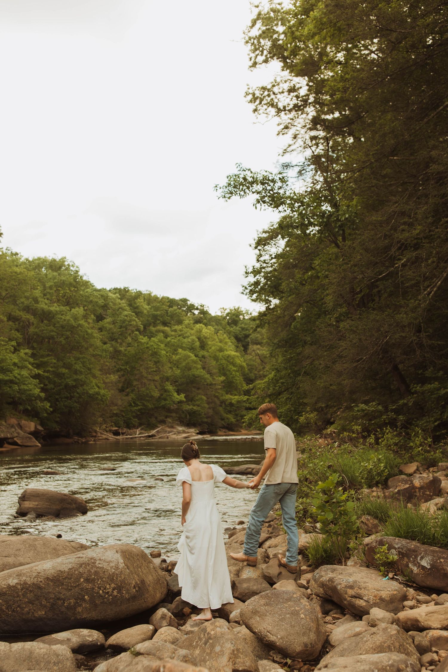 A couple explores rocky riverbanks surrounded by lush forest scenery.