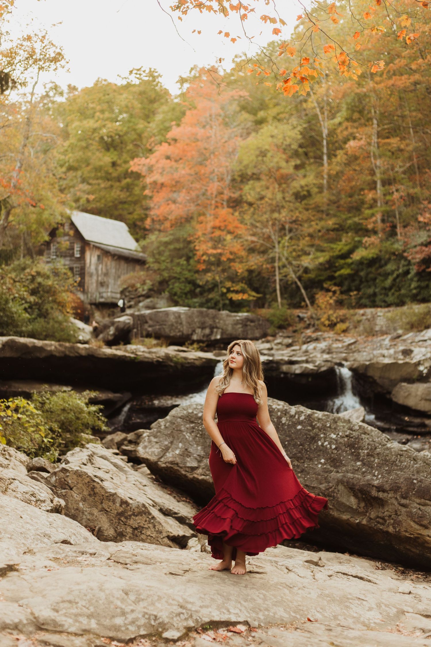 A flowing burgundy dress twirls against the backdrop of autumn trees and a rustic cabin near a rocky waterfall.