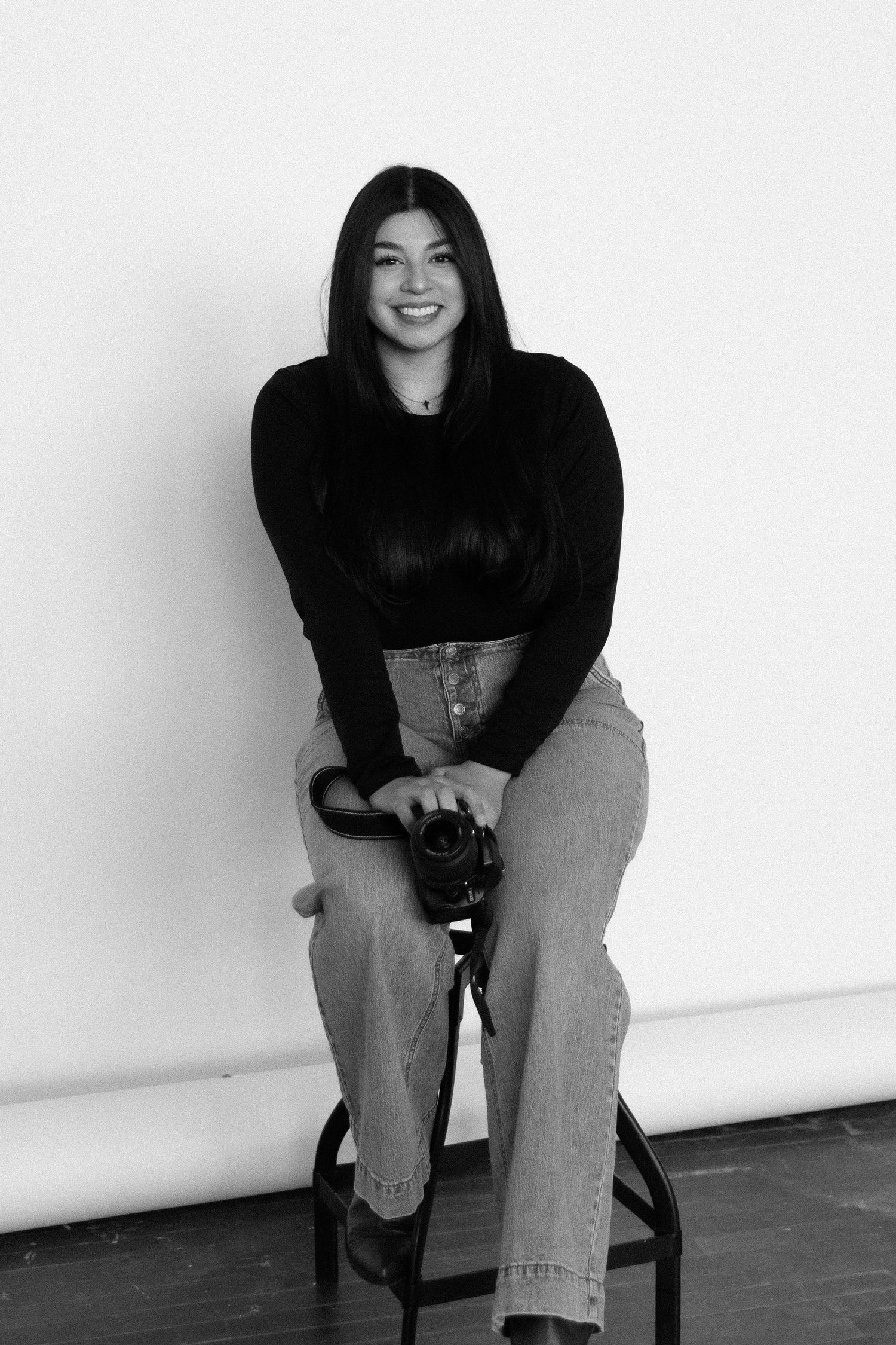 Black and white portrait of photographer sitting on stool with camera in studio setting.