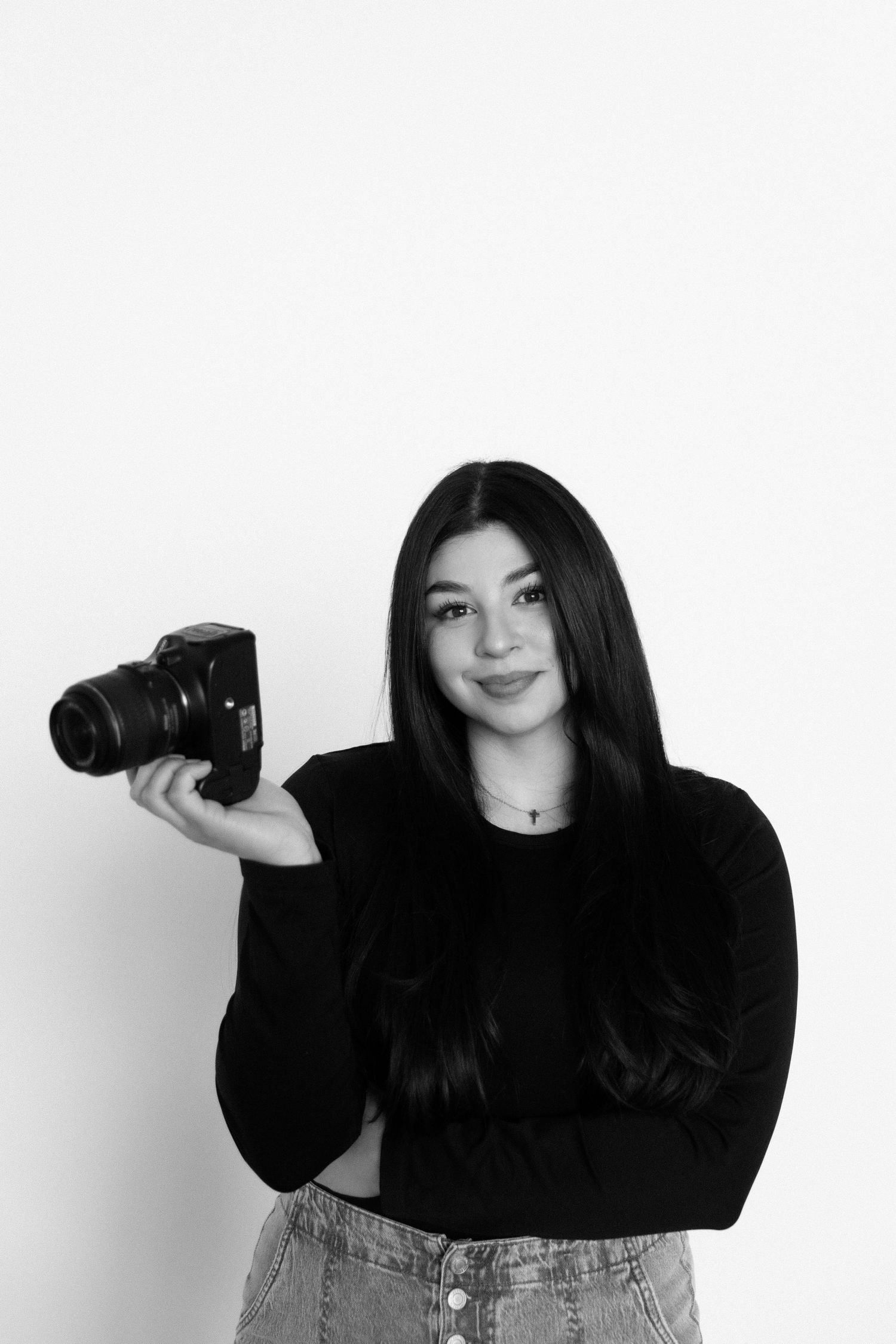 Photographer holds camera while posing in casual black sweater and jeans against white backdrop.