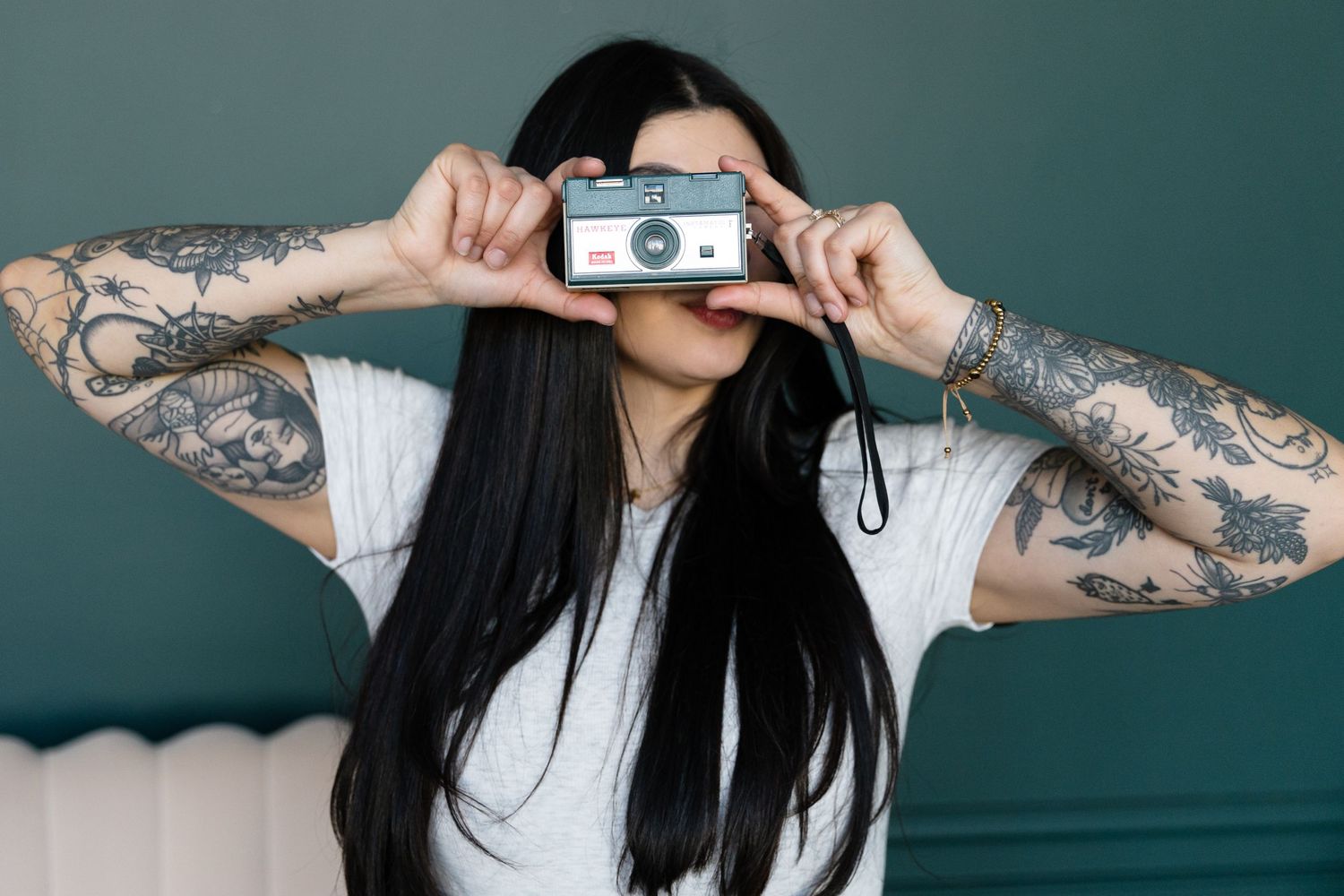 A photographer with tattooed arms holds vintage camera in front of face.
