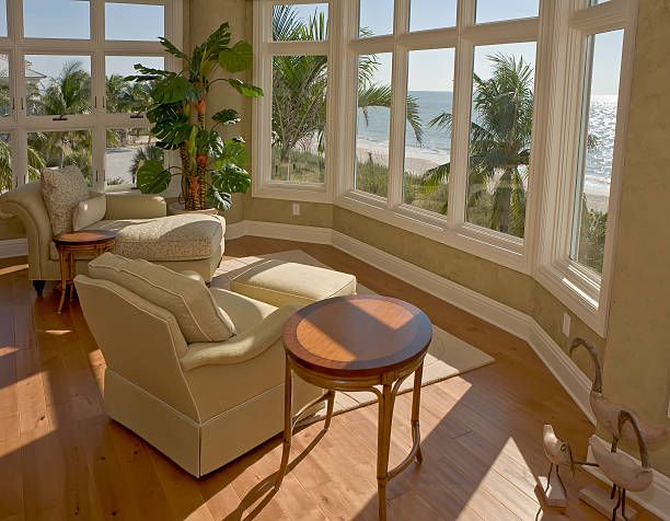 Sunlit coastal sitting area with curved windows offering panoramic ocean views and comfortable cream-colored armchairs on hardwood floors.