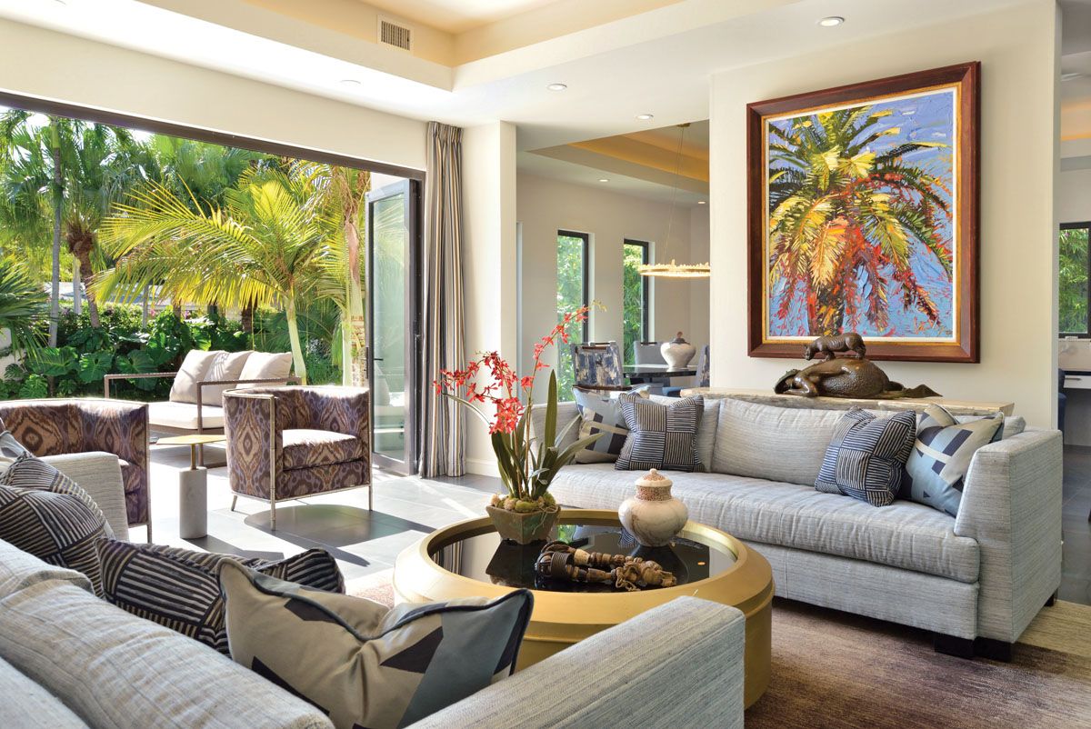 Luxurious living room with sliding glass doors opening to tropical garden, featuring grey sofas and palm tree artwork.