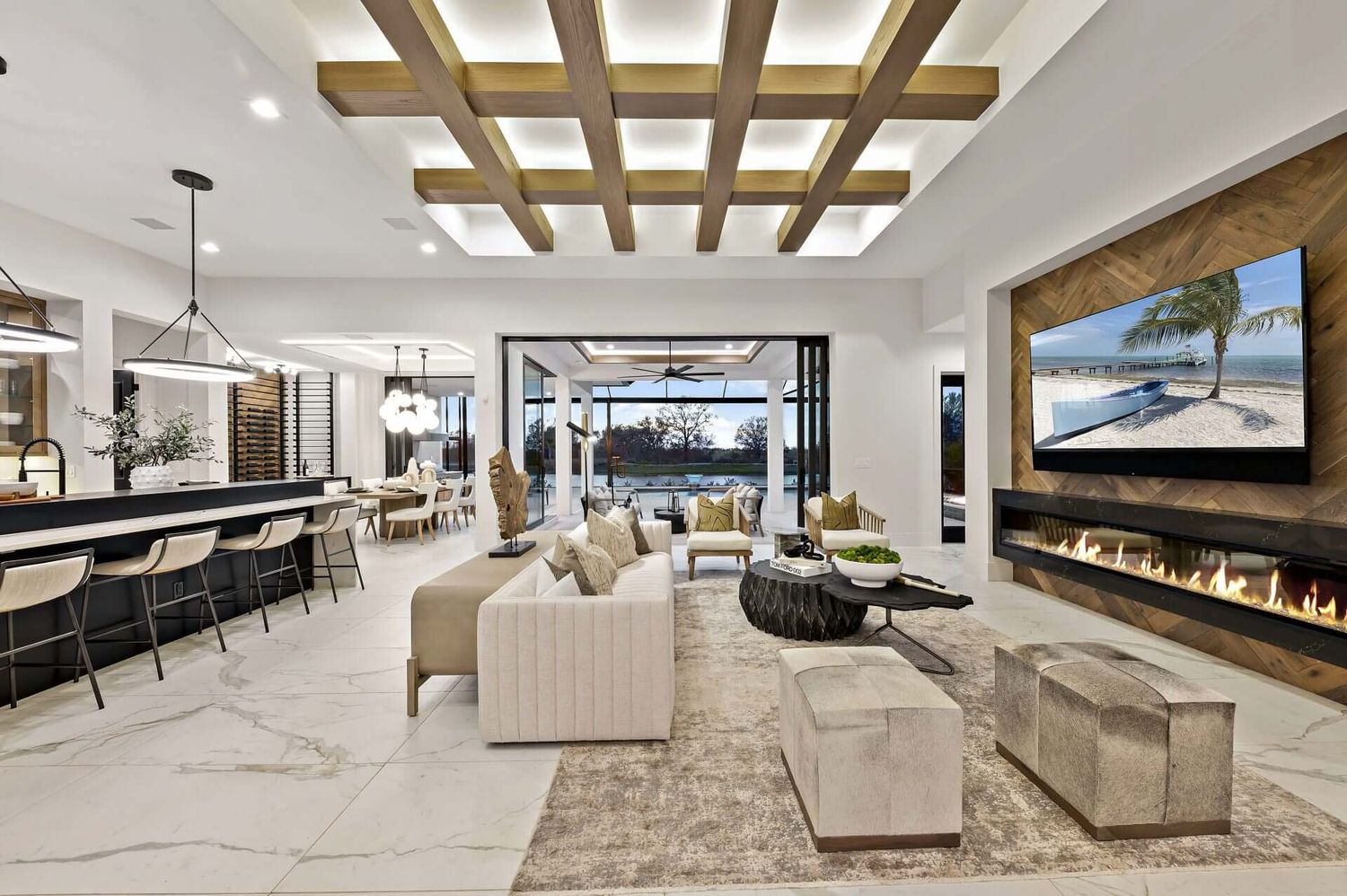 Modern open concept living space with linear fireplace, coffered ceiling, and marble floors overlooking water views.