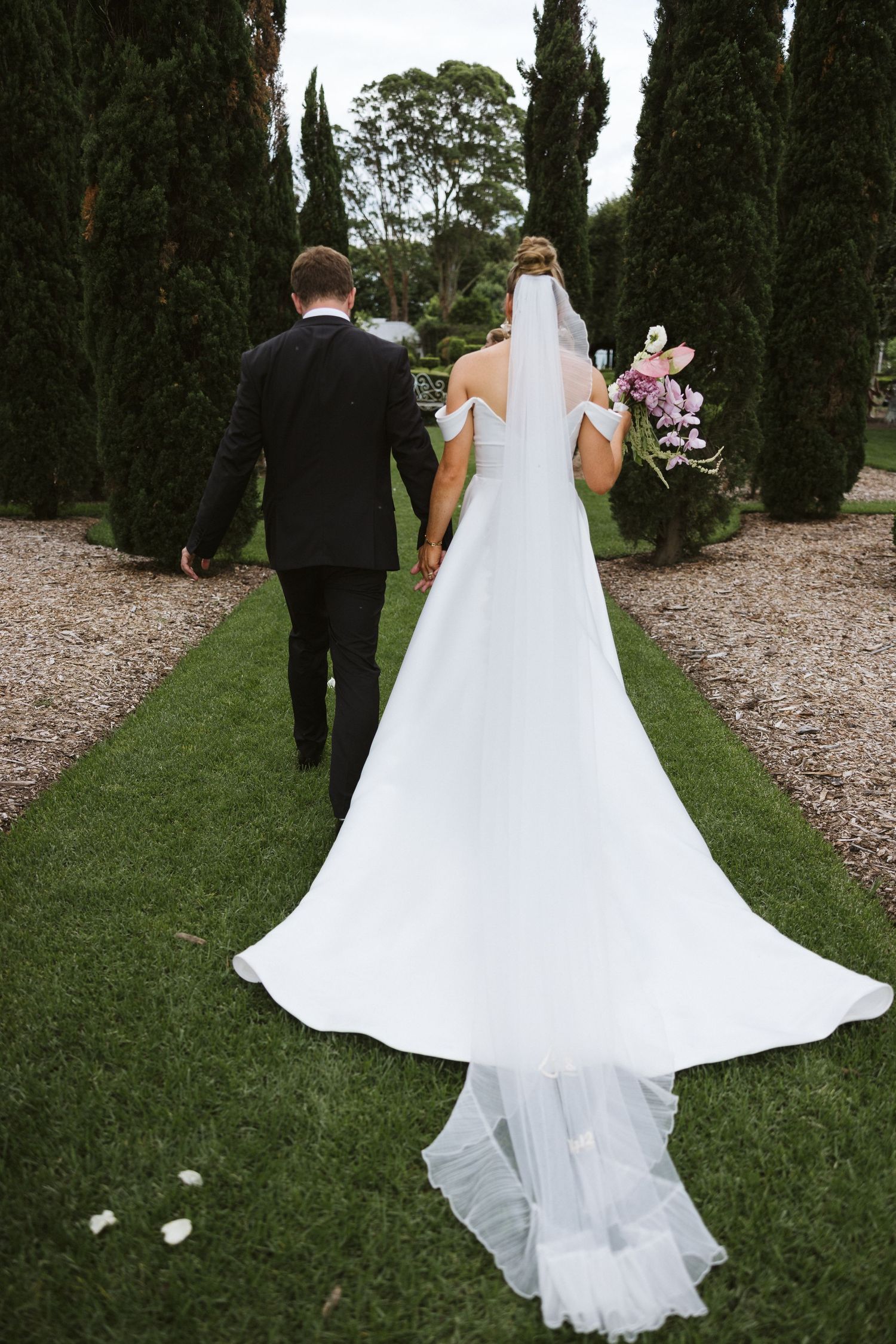 Wedding couple walking away on a cypress-lined path with flowing white train trailing behind.